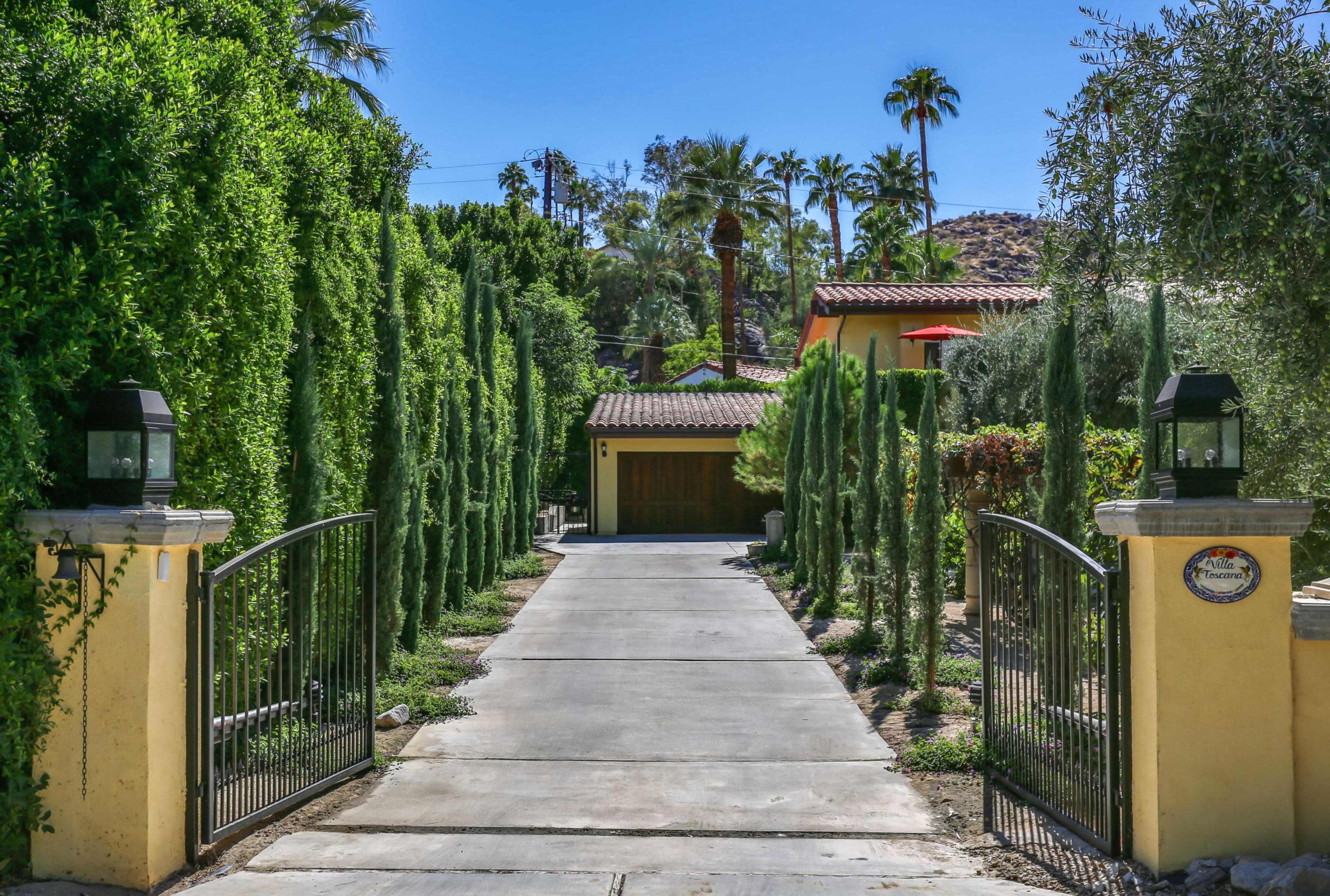 A driveway lined with tall cypress trees leads to a house surrounded by palm trees and landscaping.