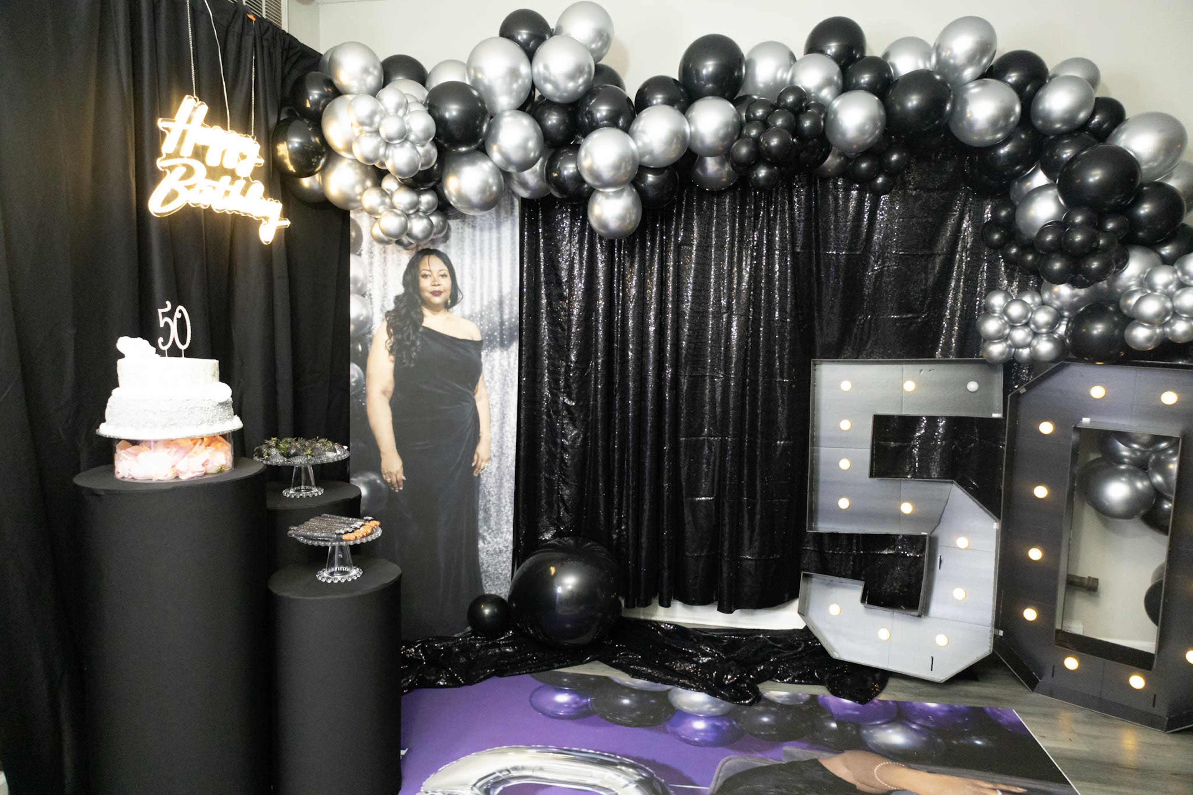The scene features a decorated party backdrop with black and silver balloons, a large "50" sign, a cake display, and a photo of a woman in an elegant dress.