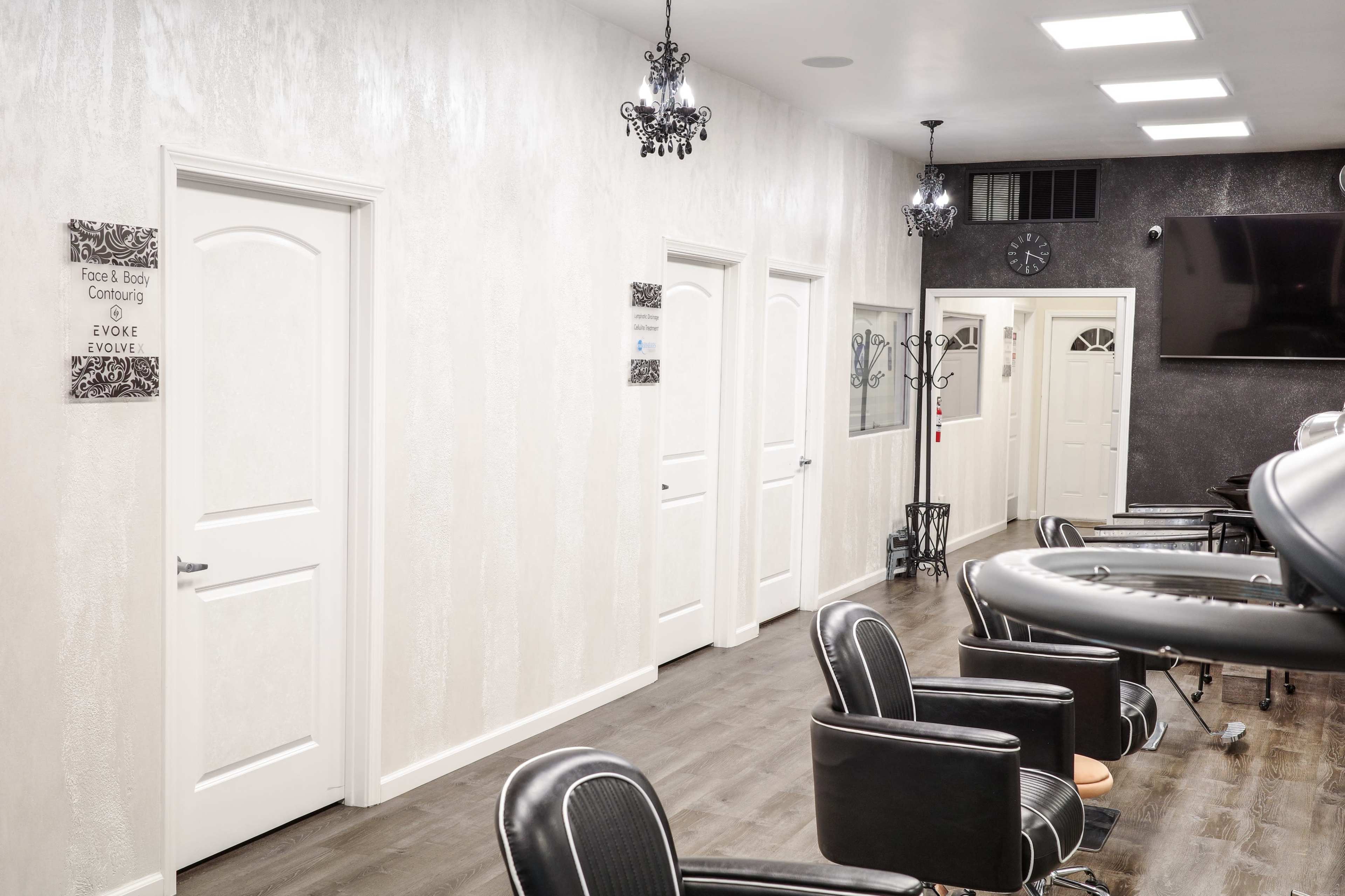 A brightly lit salon corridor features several closed doors, black salon chairs, and decorative chandeliers.