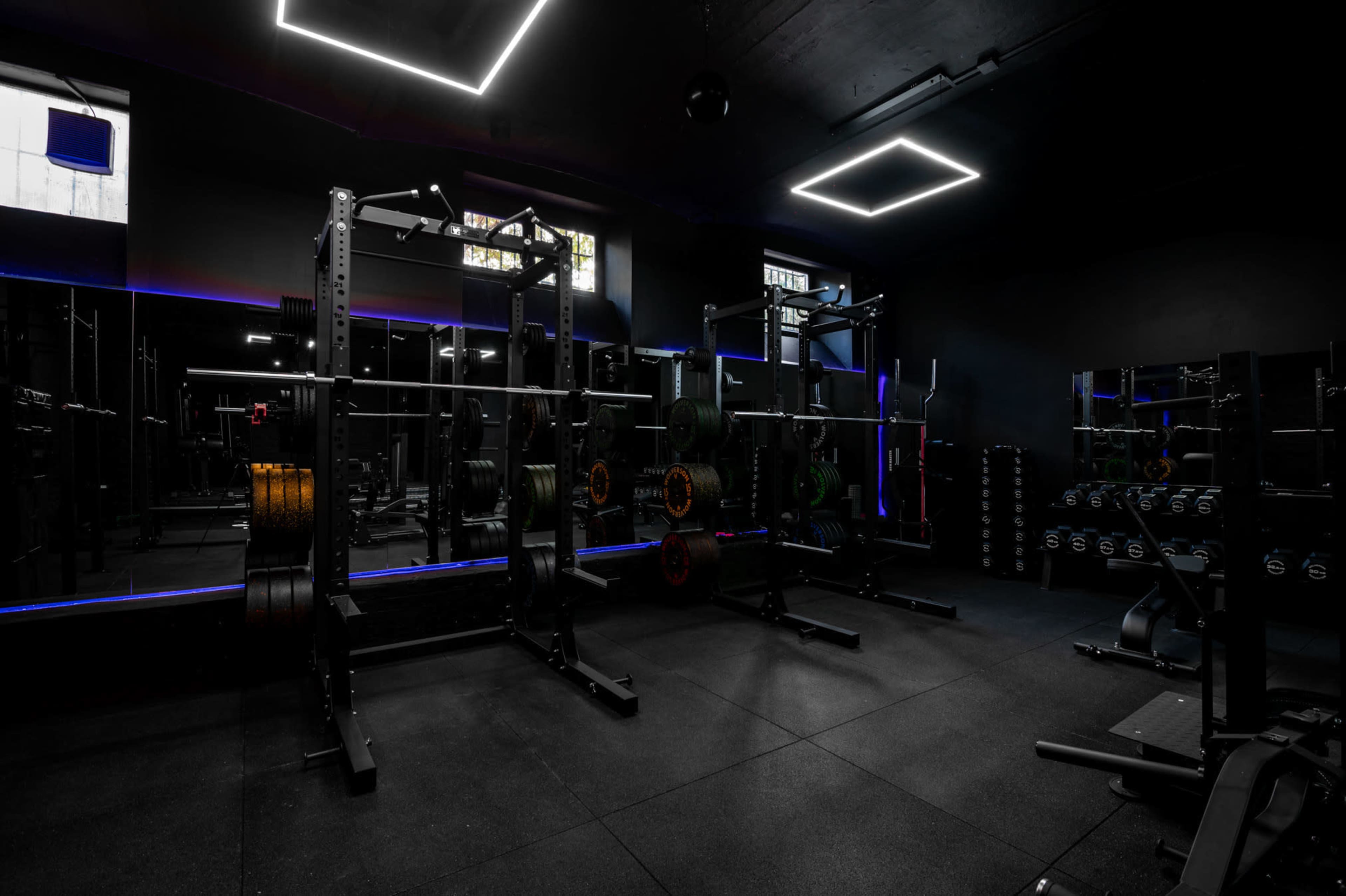 The image shows a dimly lit gym with weightlifting equipment, including squat racks and dumbbells, set against dark walls and illuminated by neon light strips.