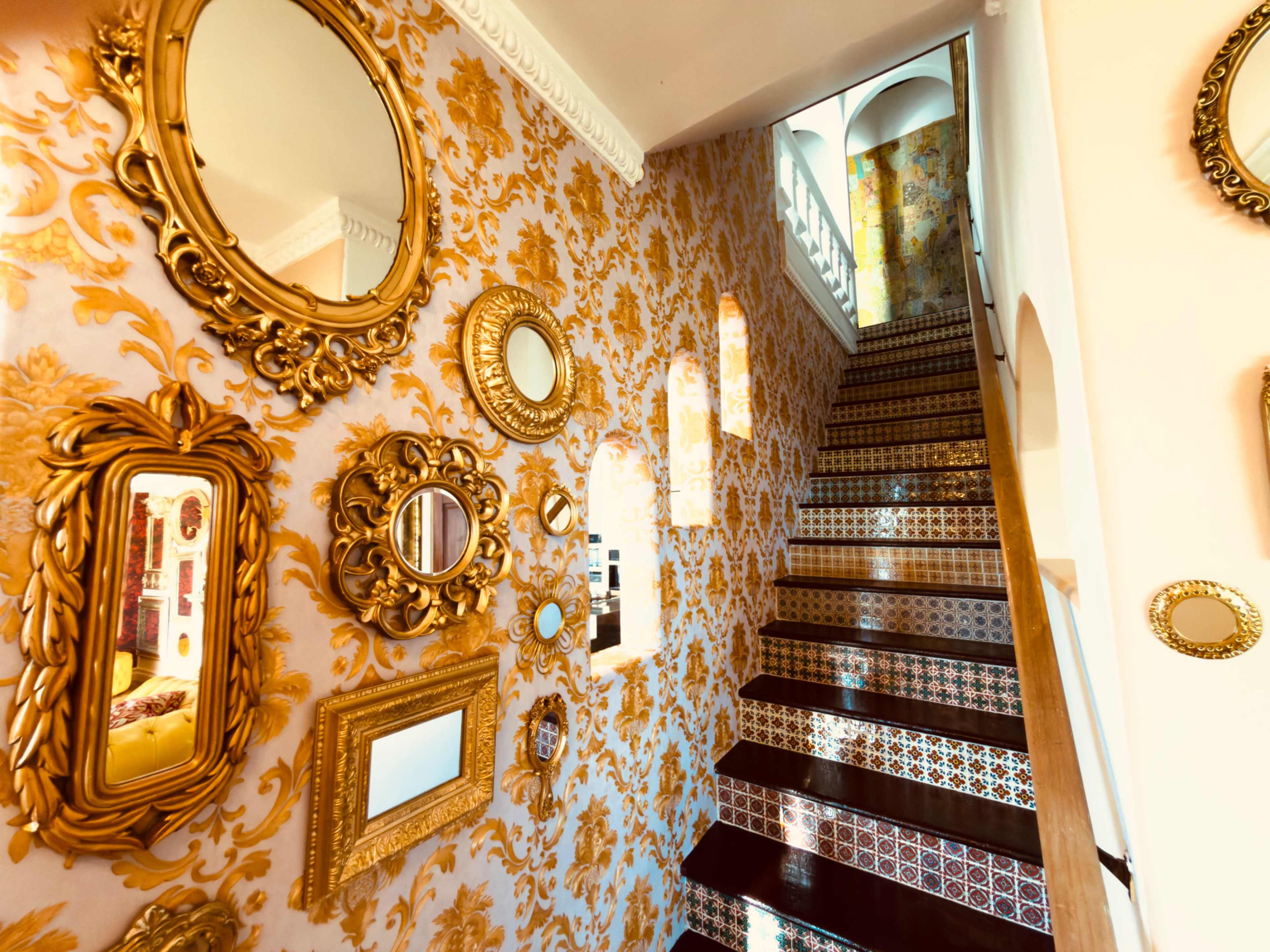 The image shows a staircase with ornate golden mirrors and patterned wallpaper, leading up to a brightly lit area.