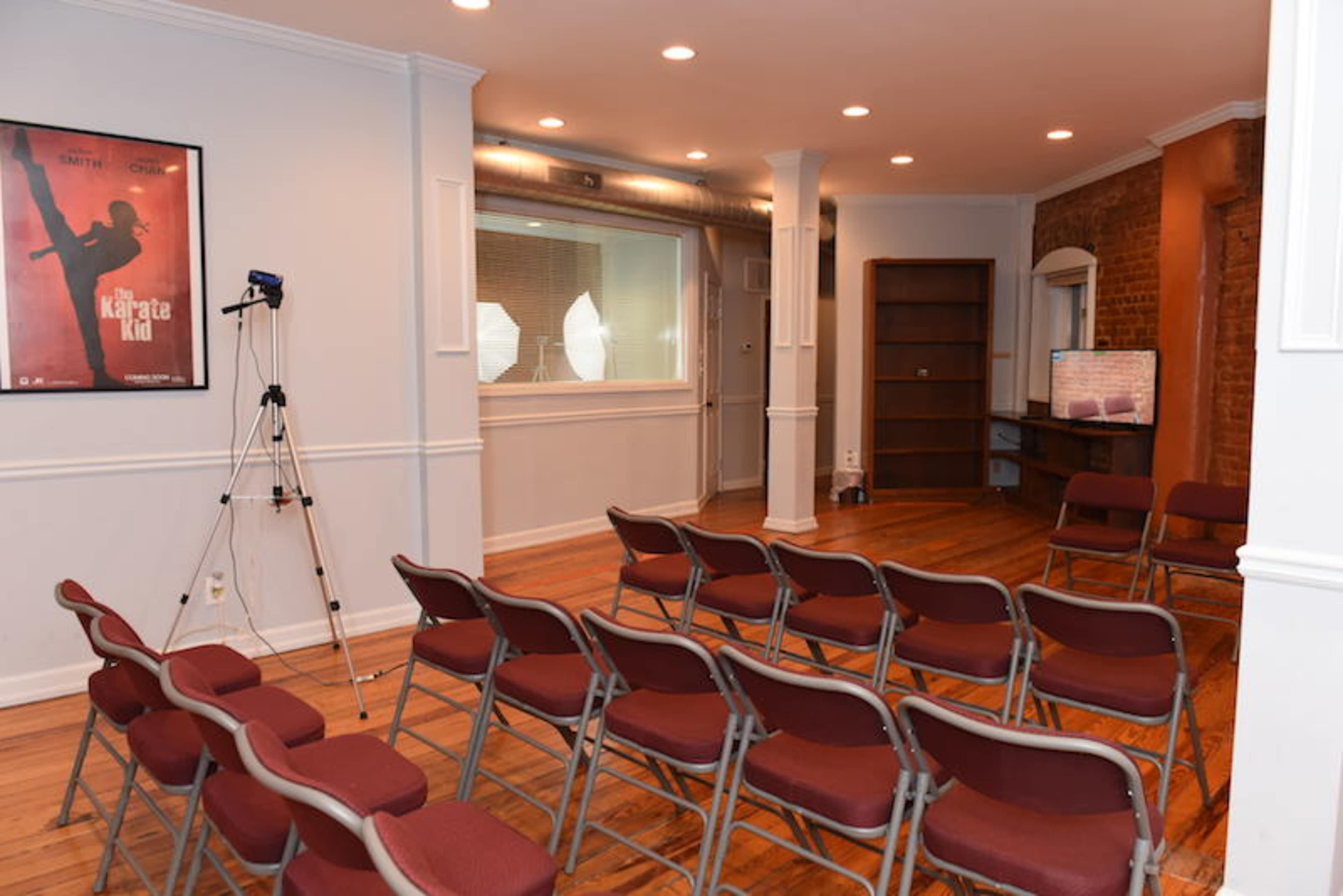 A room with rows of red chairs facing a glass-paneled room, and a poster on the wall next to a camera tripod.