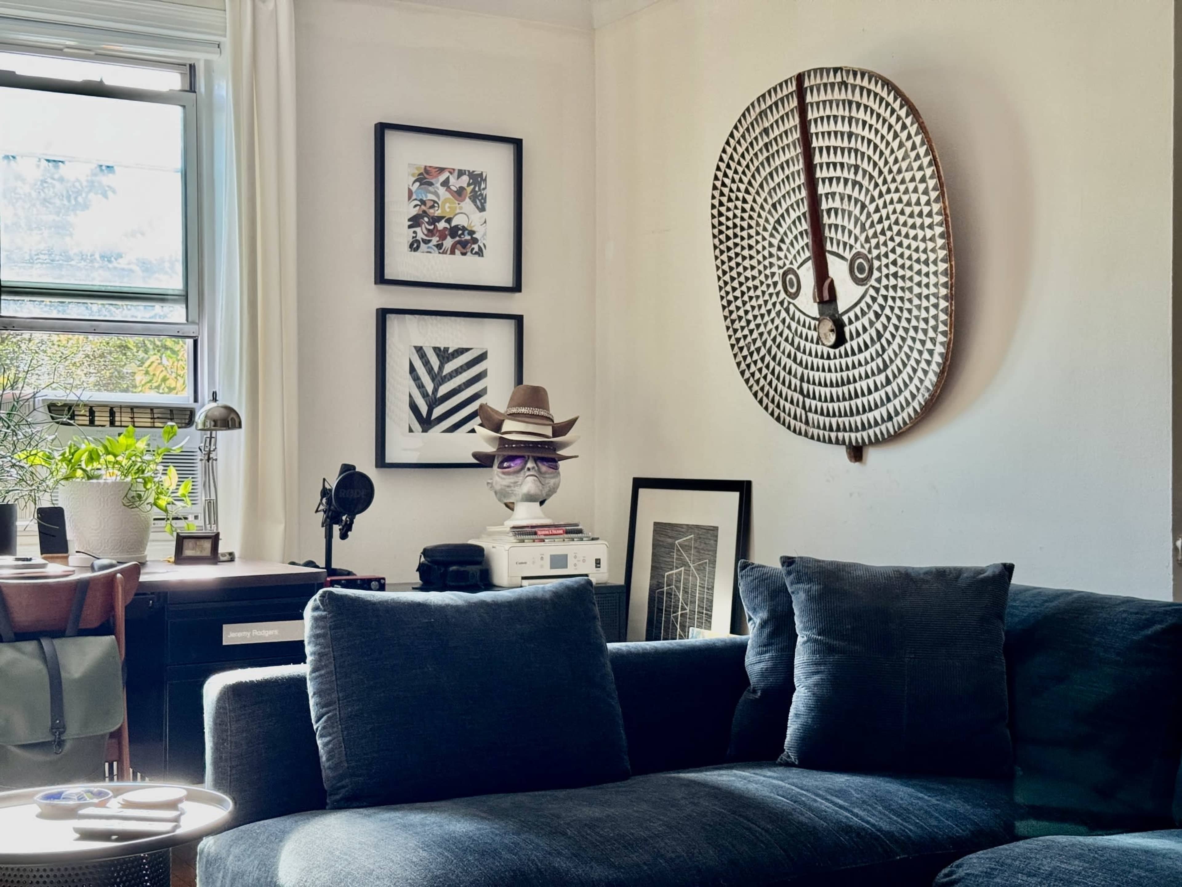 The image shows a living room with a dark blue couch, an assortment of framed art on the walls, and a decorative mask hanging above the couch.