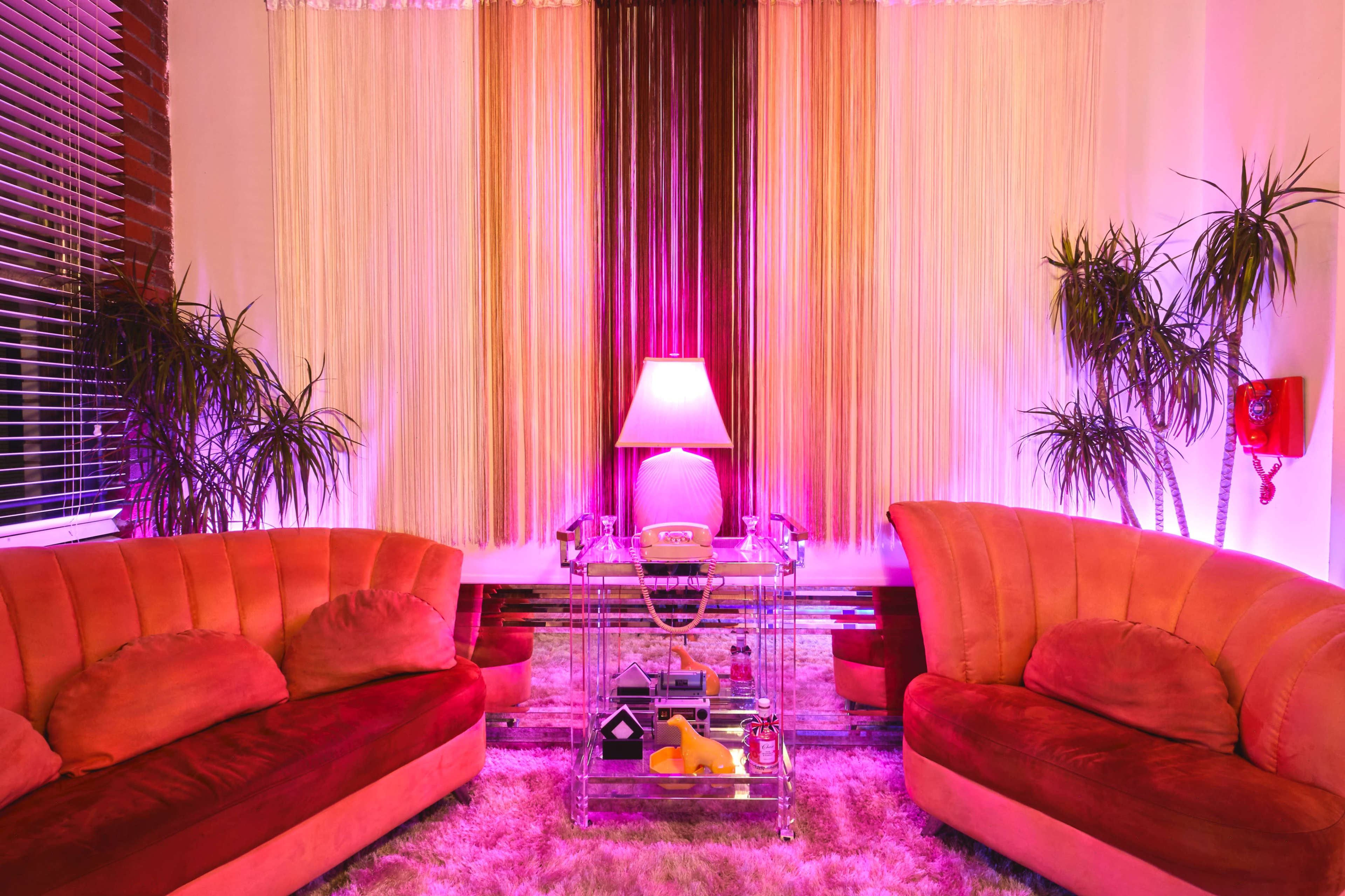 A brightly lit retro living room features two curved, pink sofas, a glass coffee table, and a floor lamp, surrounded by potted plants and colorful curtains.