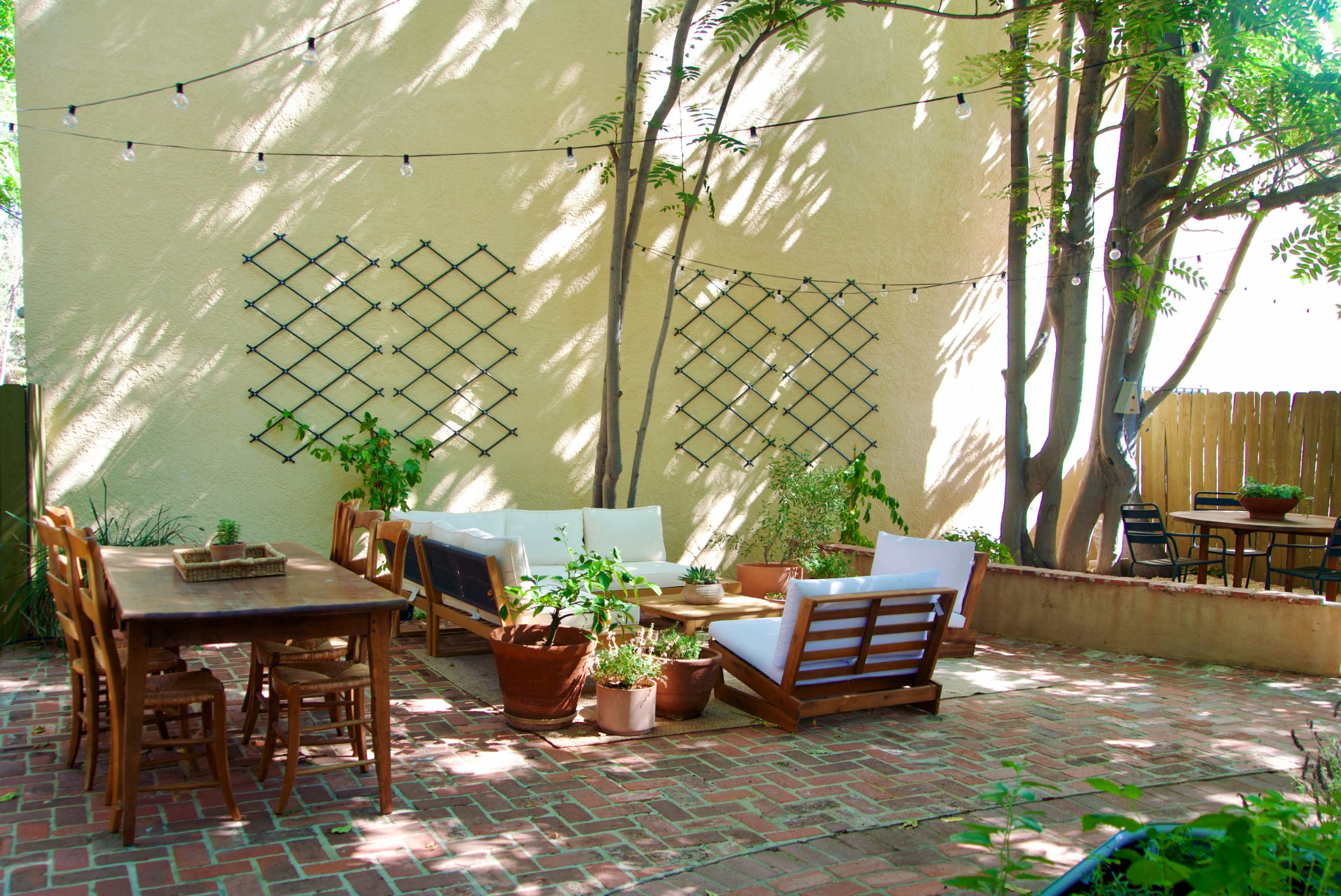 The image shows a brick patio area with seating, tables, and plants under strings of lights adjacent to a wall.
