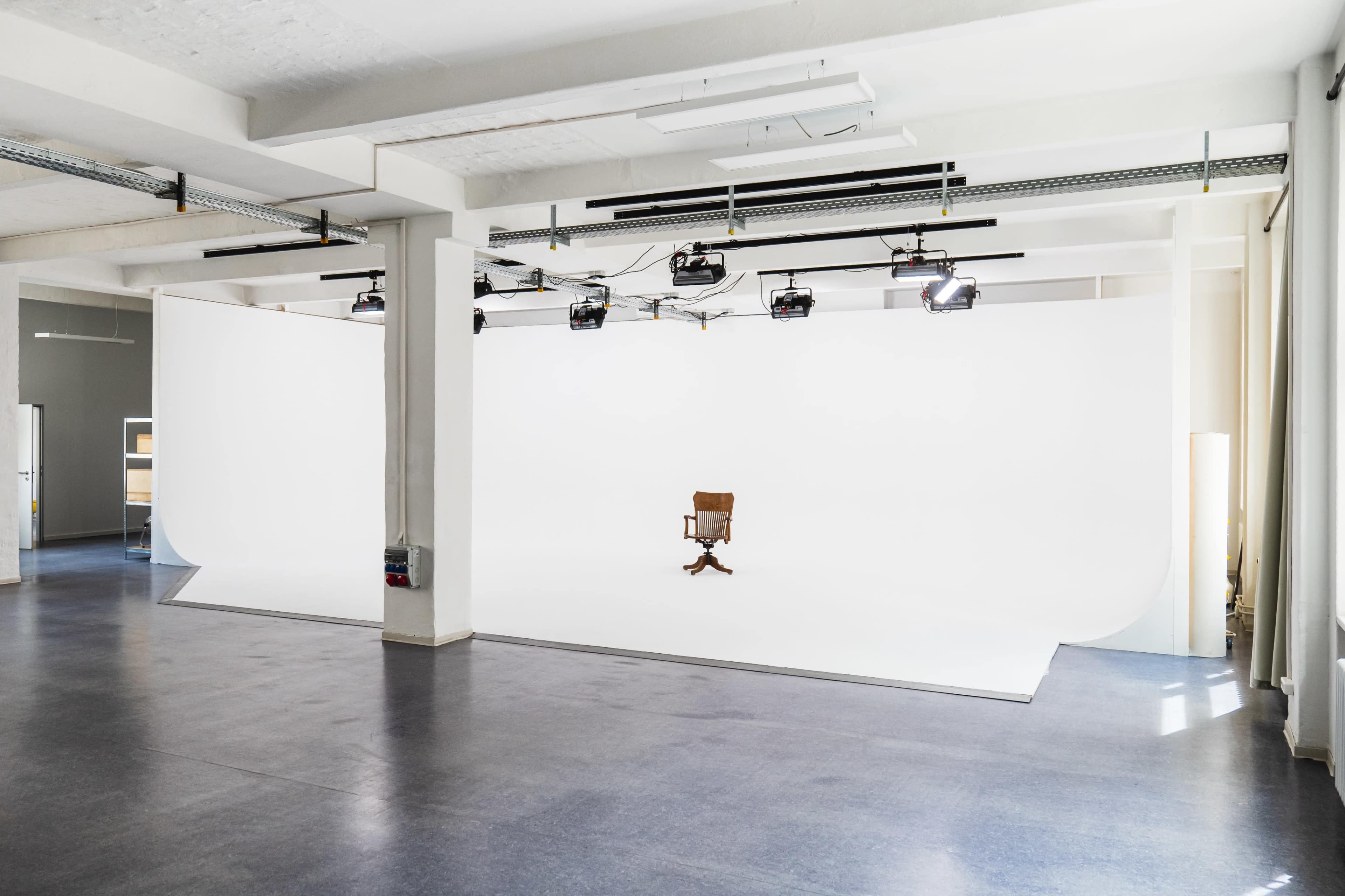 A spacious studio with a white backdrop, spotlights, and a single wooden chair positioned in the center.