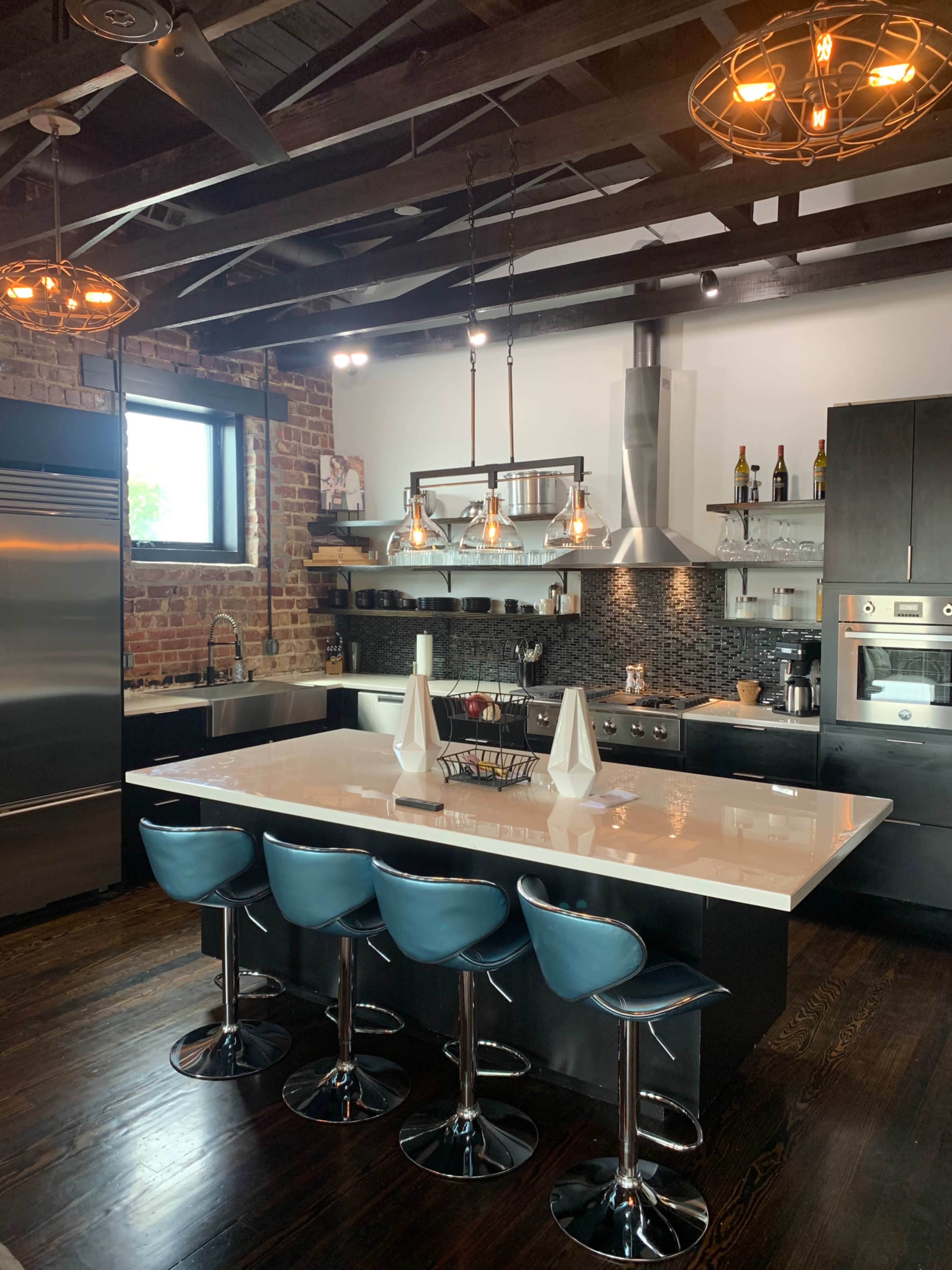 A modern kitchen features a large island with blue bar stools, exposed brick walls, pendant lighting, and stainless steel appliances.
