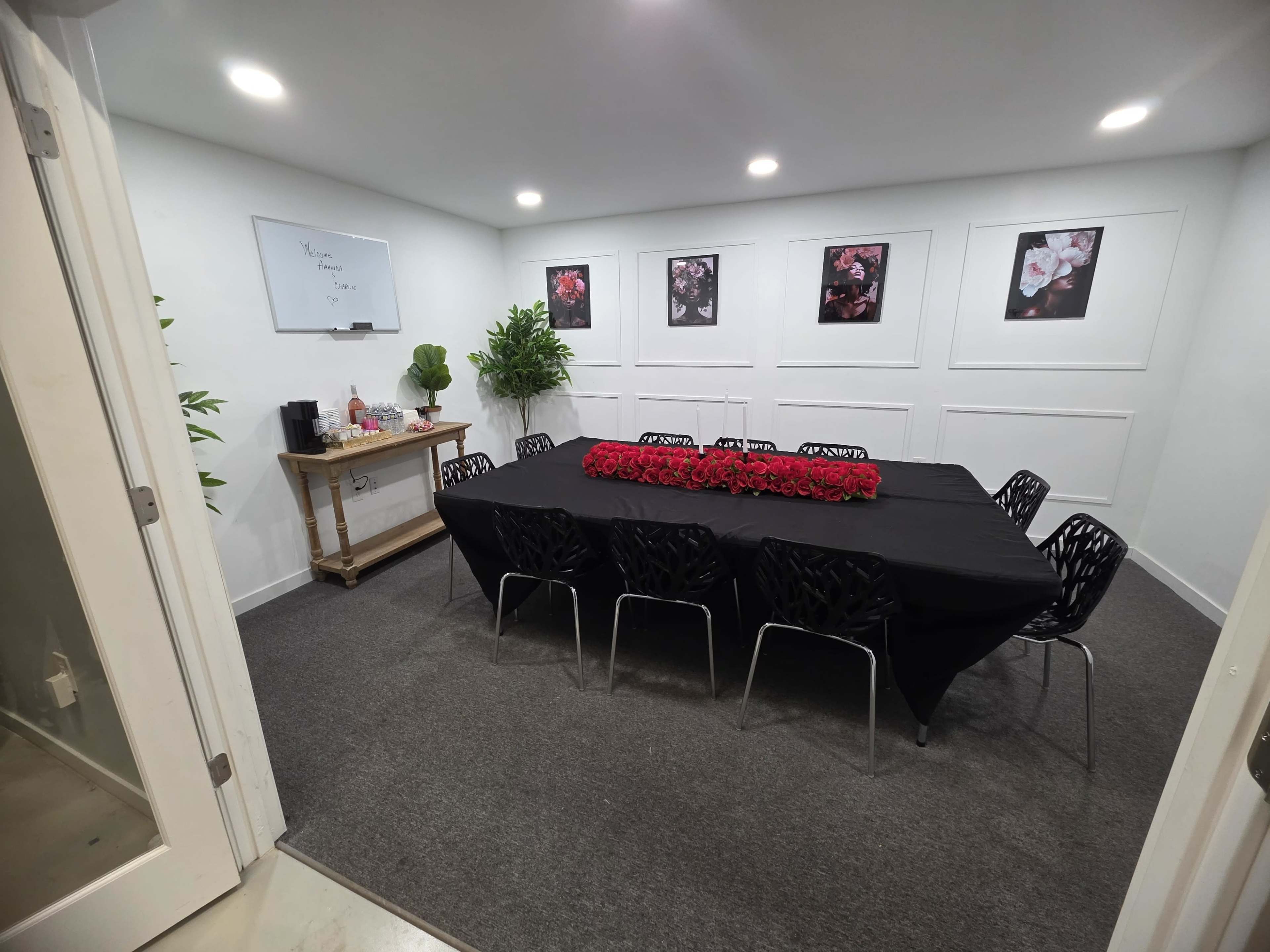 A meeting room features a long table covered with a black cloth, surrounded by chairs, and adorned with red flowers, with plants and framed photographs on the walls.