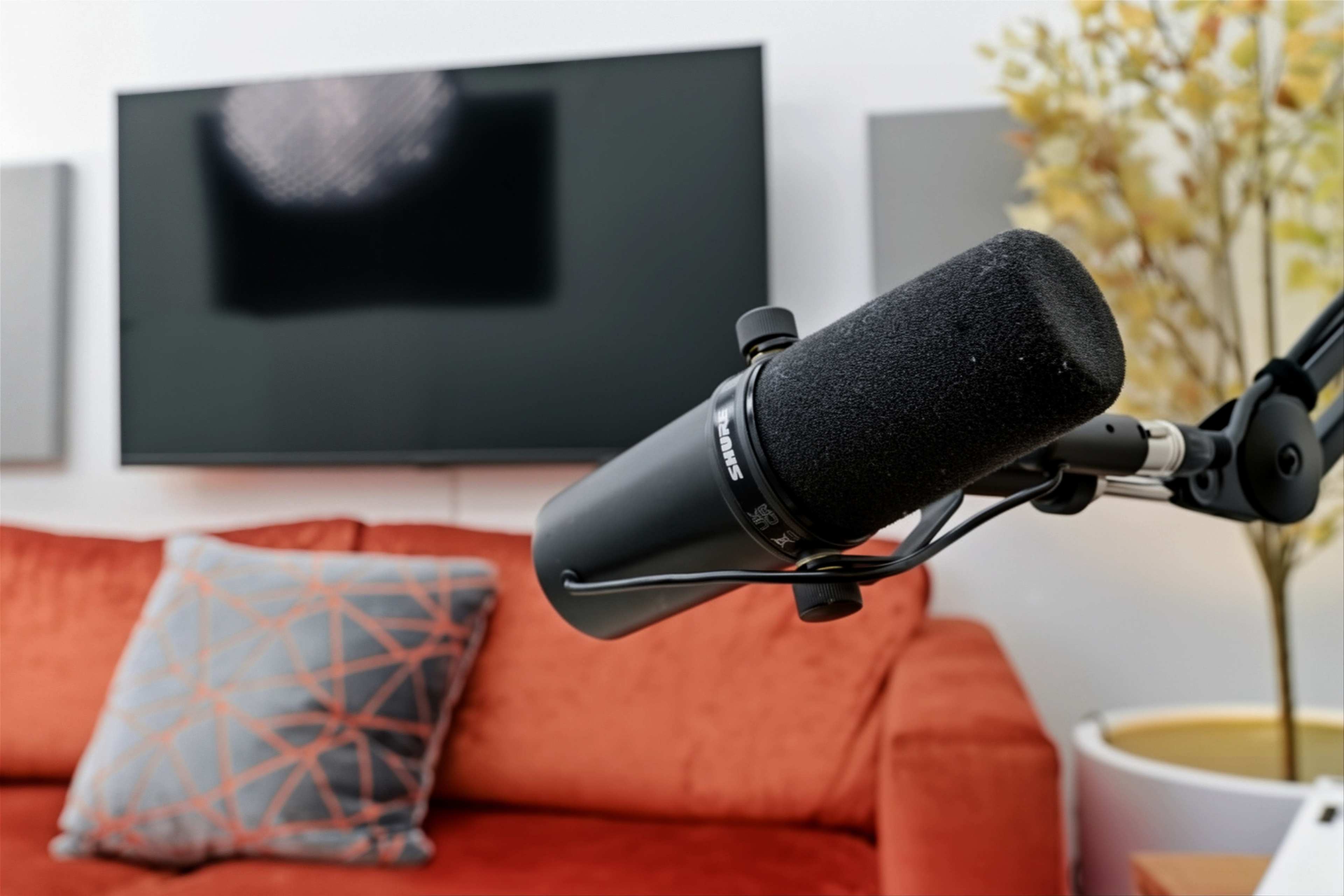 A close-up of a microphone on a stand in front of a television and an orange couch with a decorative pillow.