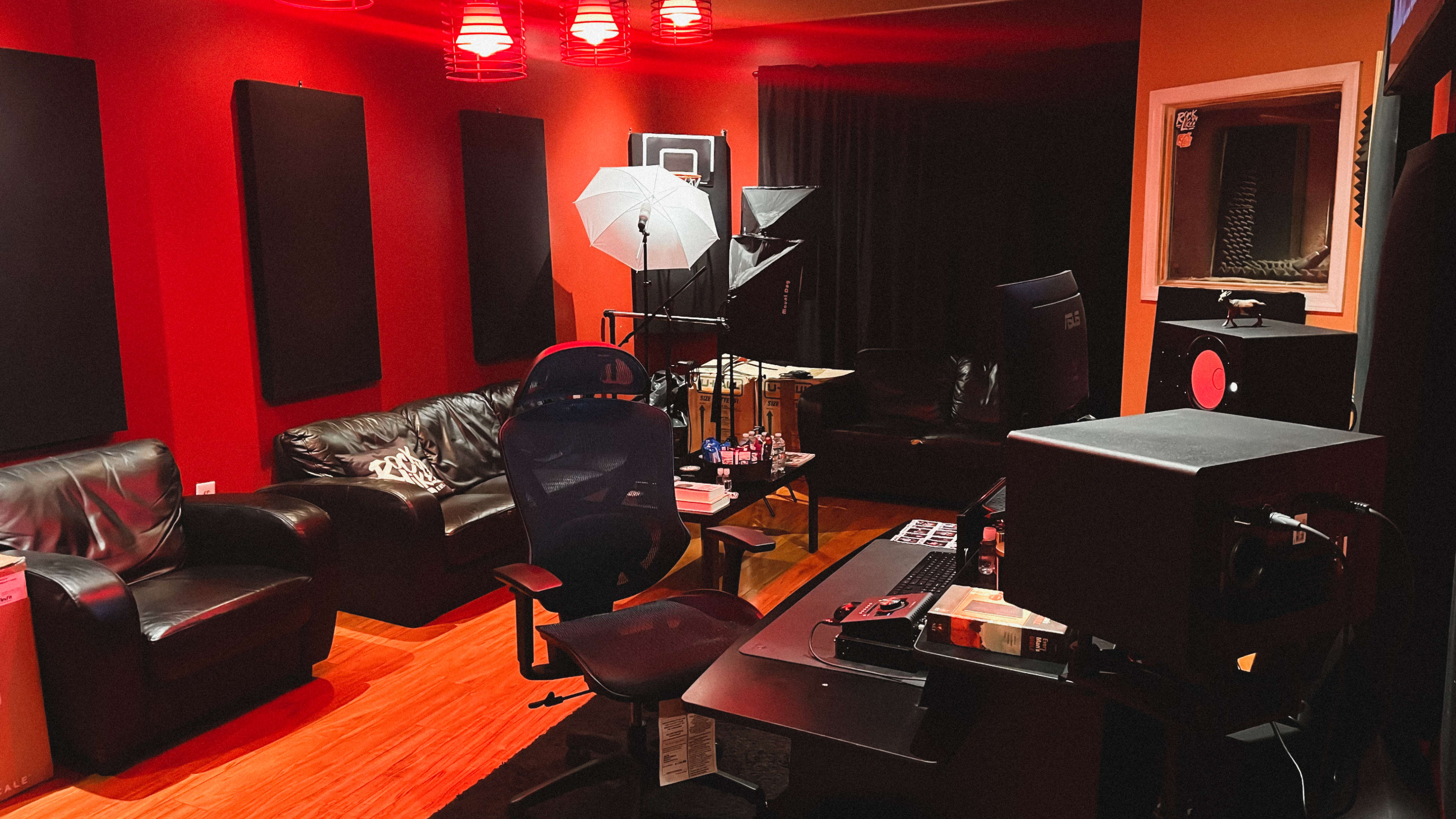 The image shows a recording studio with black leather furniture, red walls, professional lighting equipment, and a computer desk.