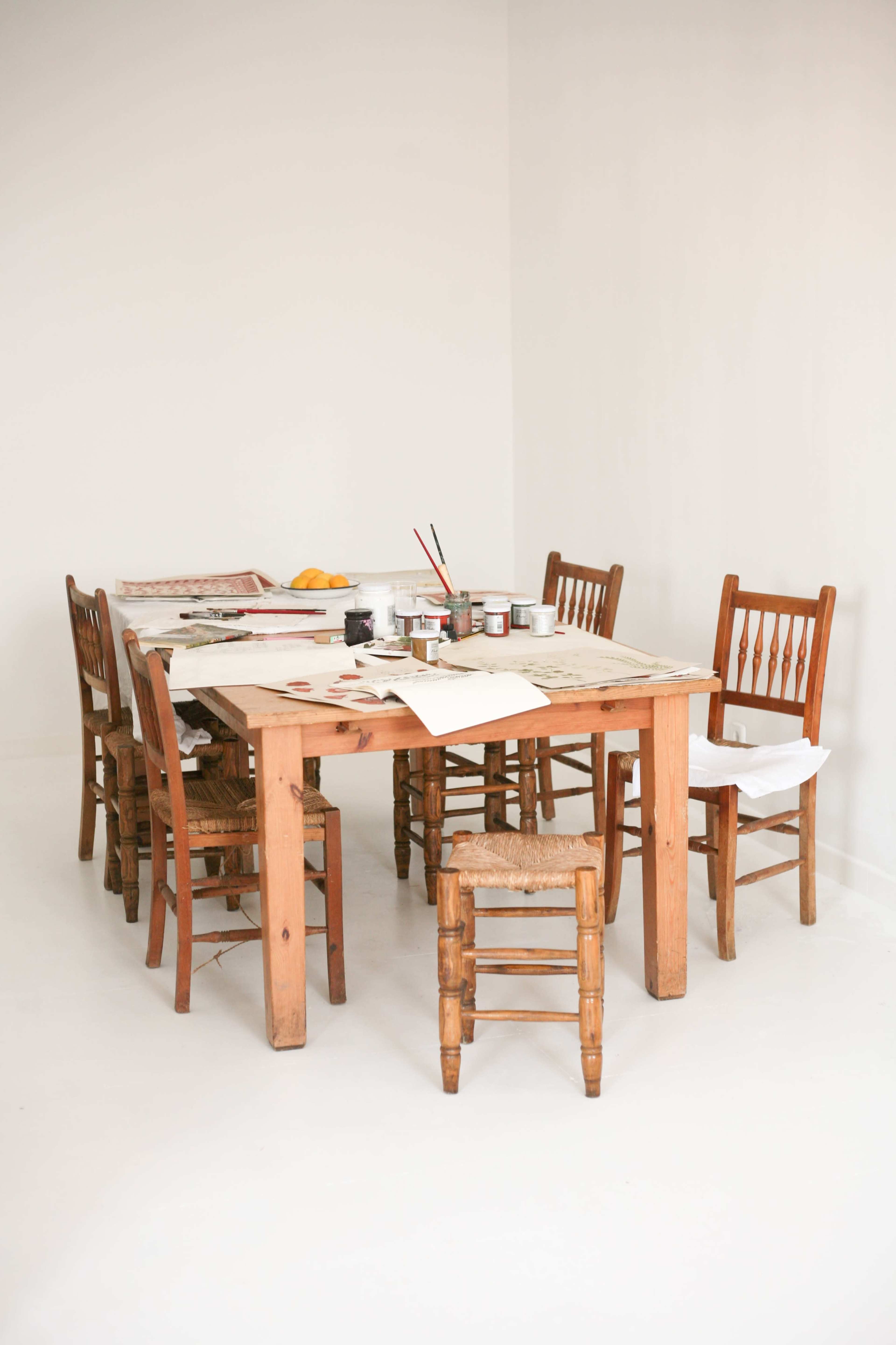 A wooden table surrounded by several chairs is set in a minimalist room, with scattered art supplies and papers on the table.