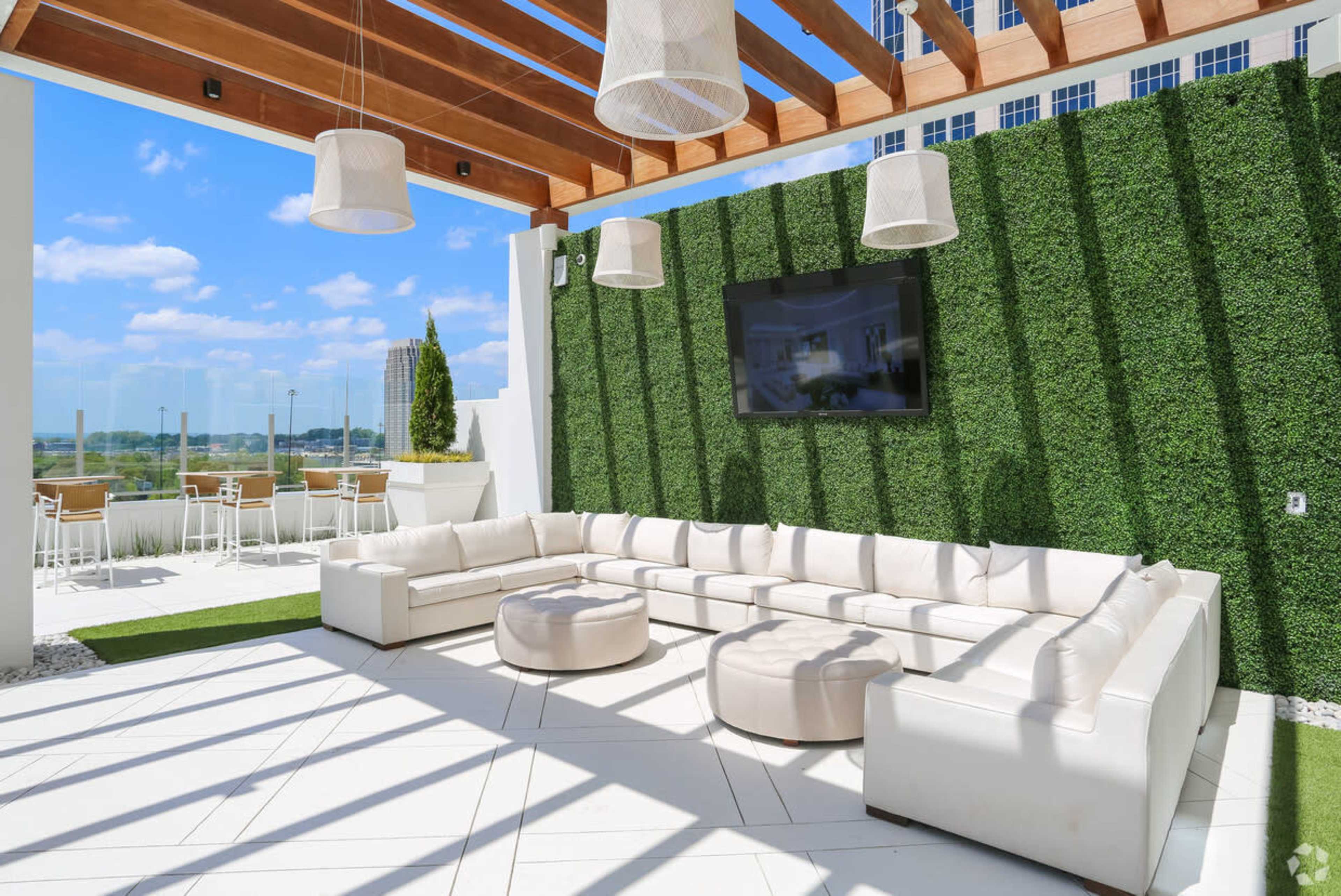 The image shows a modern outdoor lounge area with a large white sectional sofa, round seating, and a wall of greenery under a pergola with hanging light fixtures and a television.