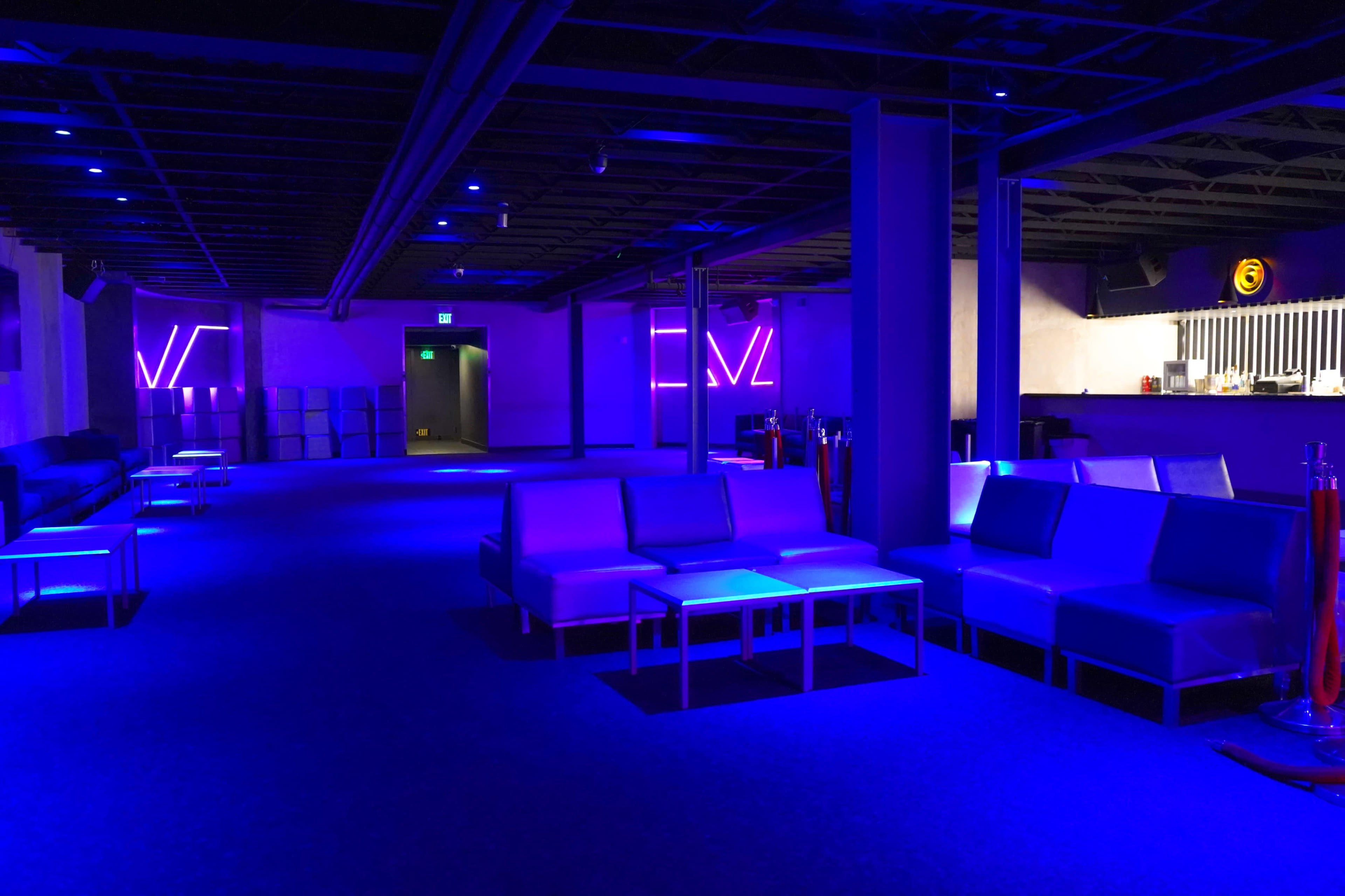 The image shows a dimly lit lounge area with blue lighting, featuring several white couches and a bar in the background.