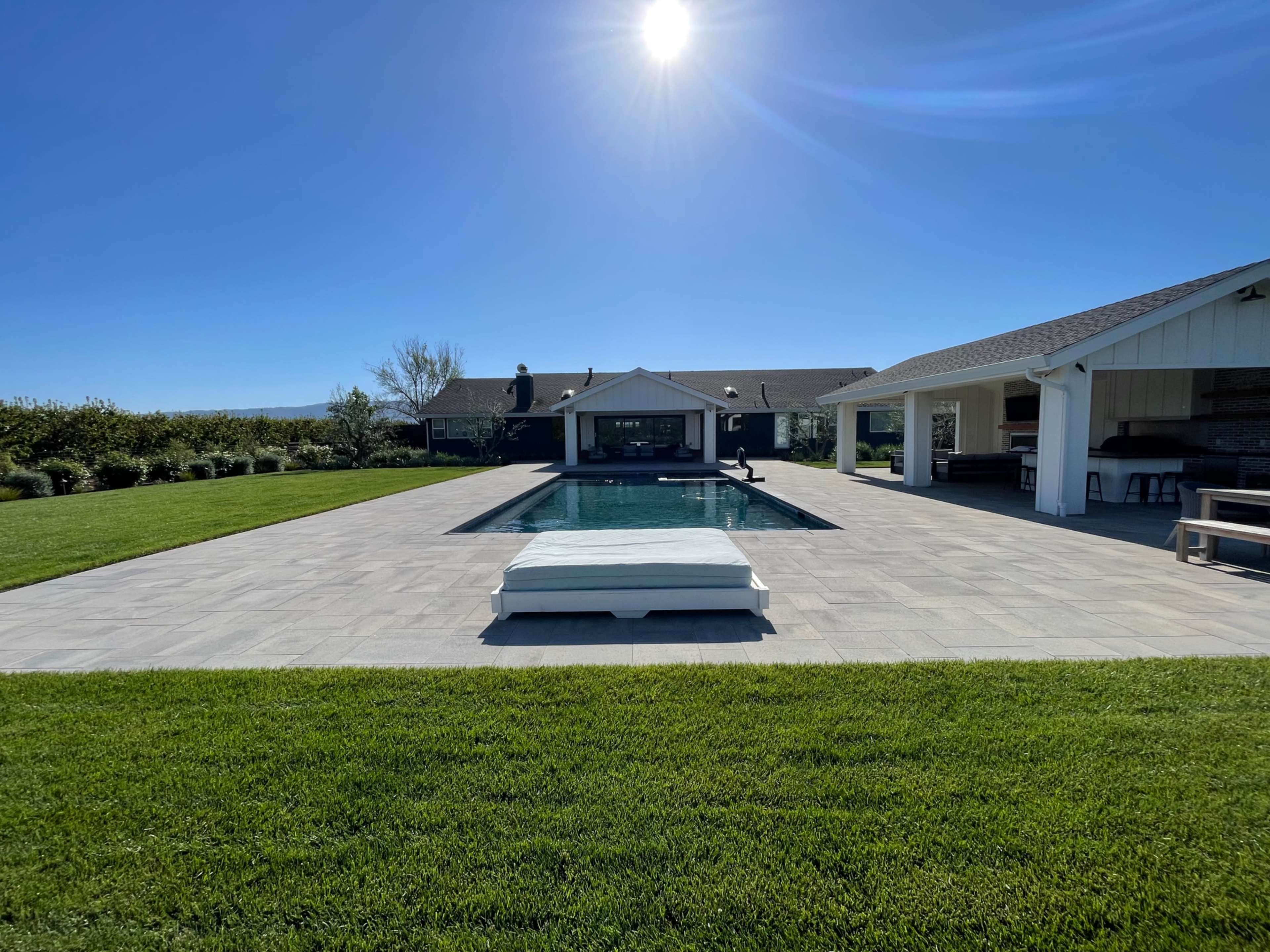 A rectangular swimming pool is surrounded by a paved patio and a grassy lawn, with a house and an outdoor kitchen visible in the background under a clear blue sky.