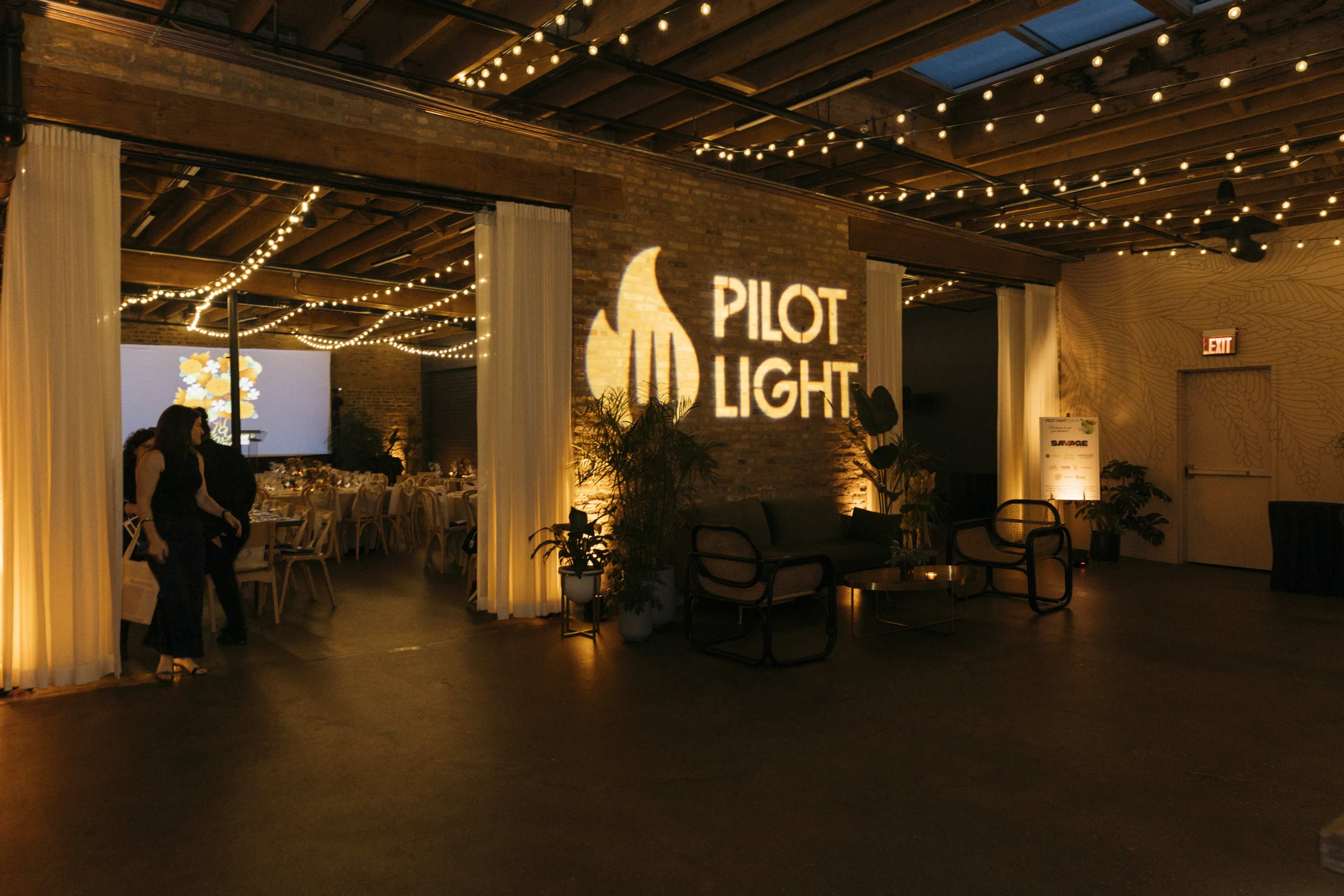 The image shows an event space with string lights, a projected logo reading "PILOT LIGHT," and arranged tables and chairs for a gathering.