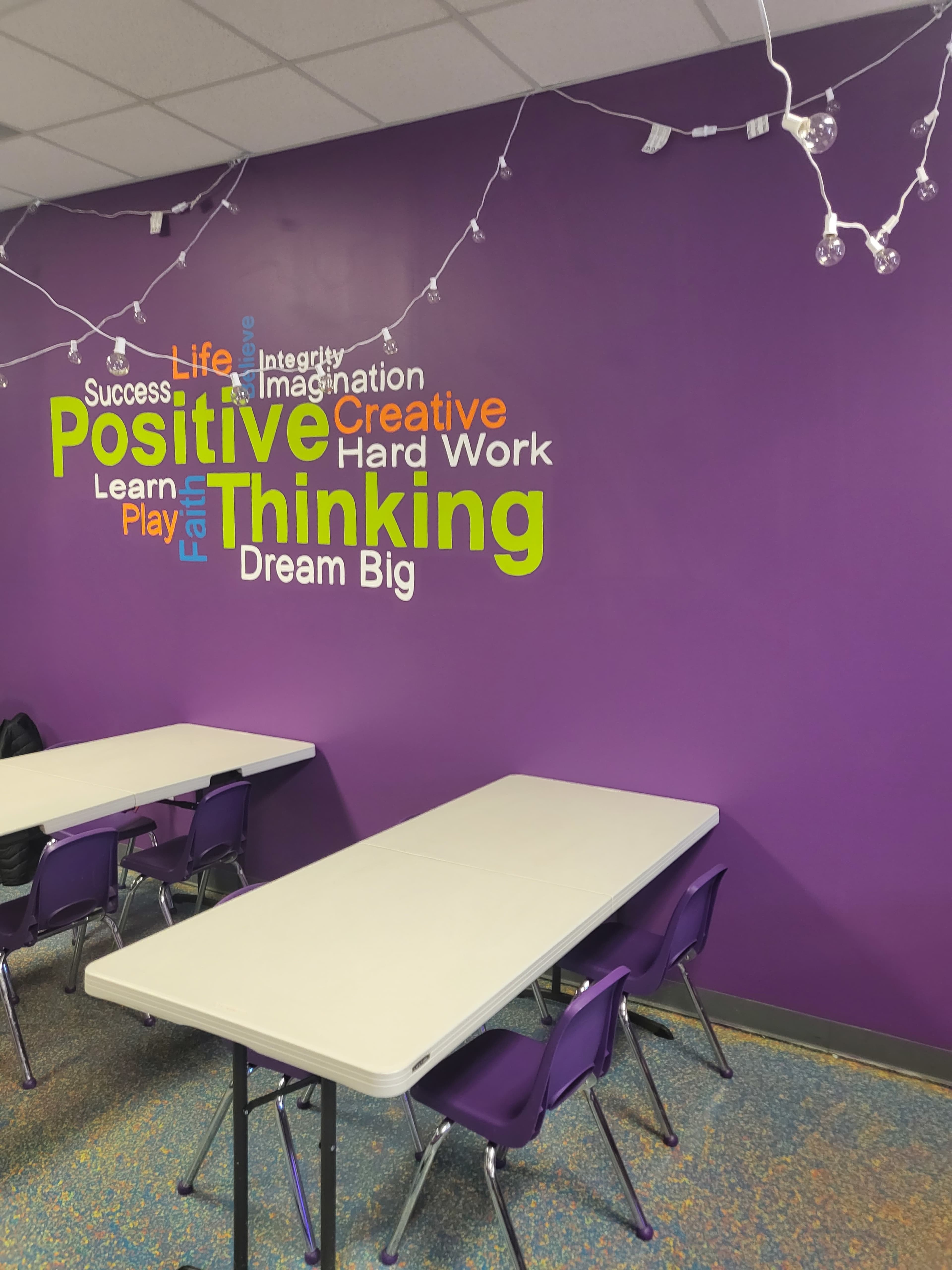 A room with purple walls featuring a word cloud of motivational phrases, alongside several tables and chairs set up for use.