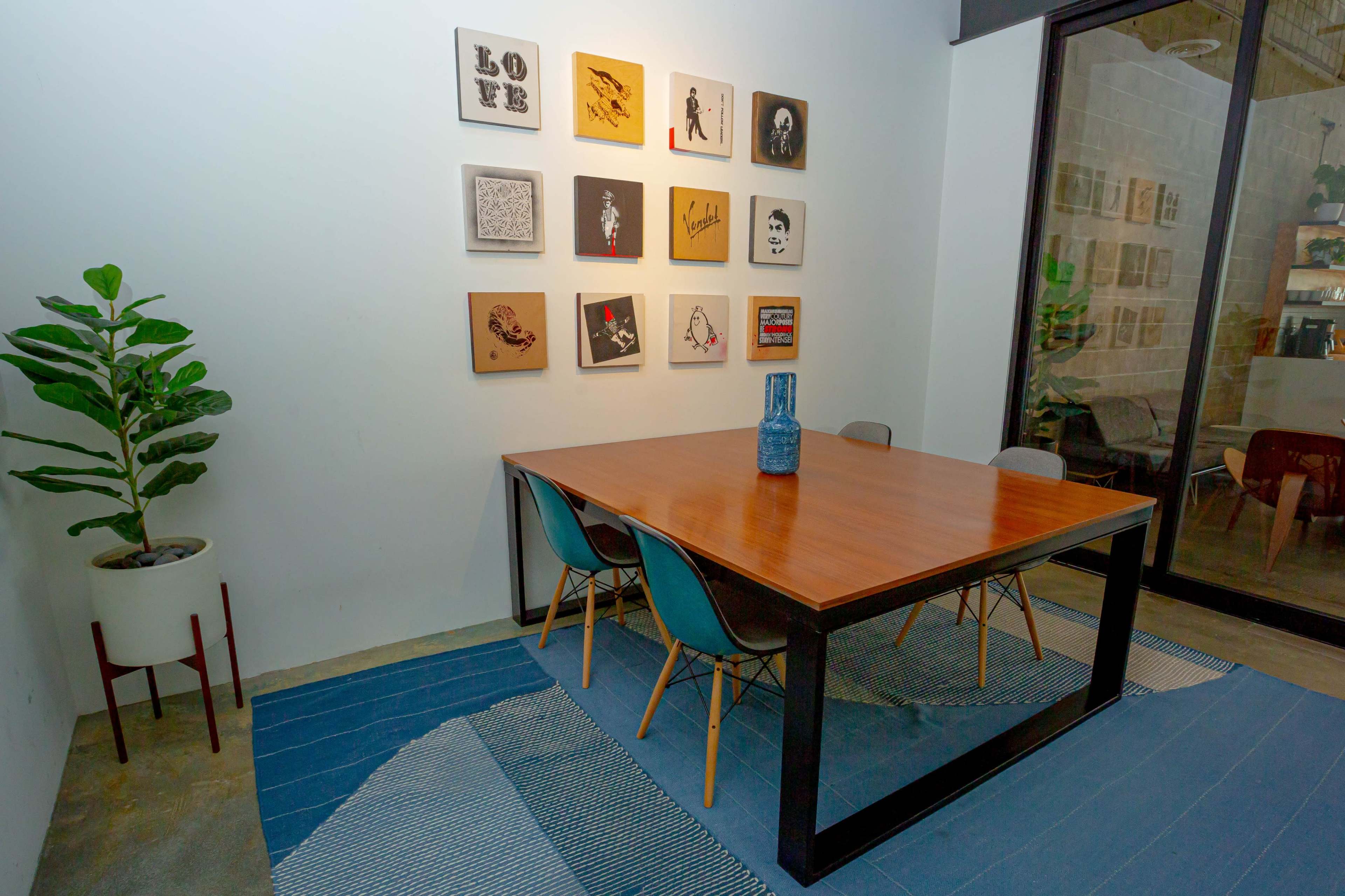 A wooden table with four chairs is set beneath a wall displaying various framed artworks in a well-lit room with a potted plant in the corner.