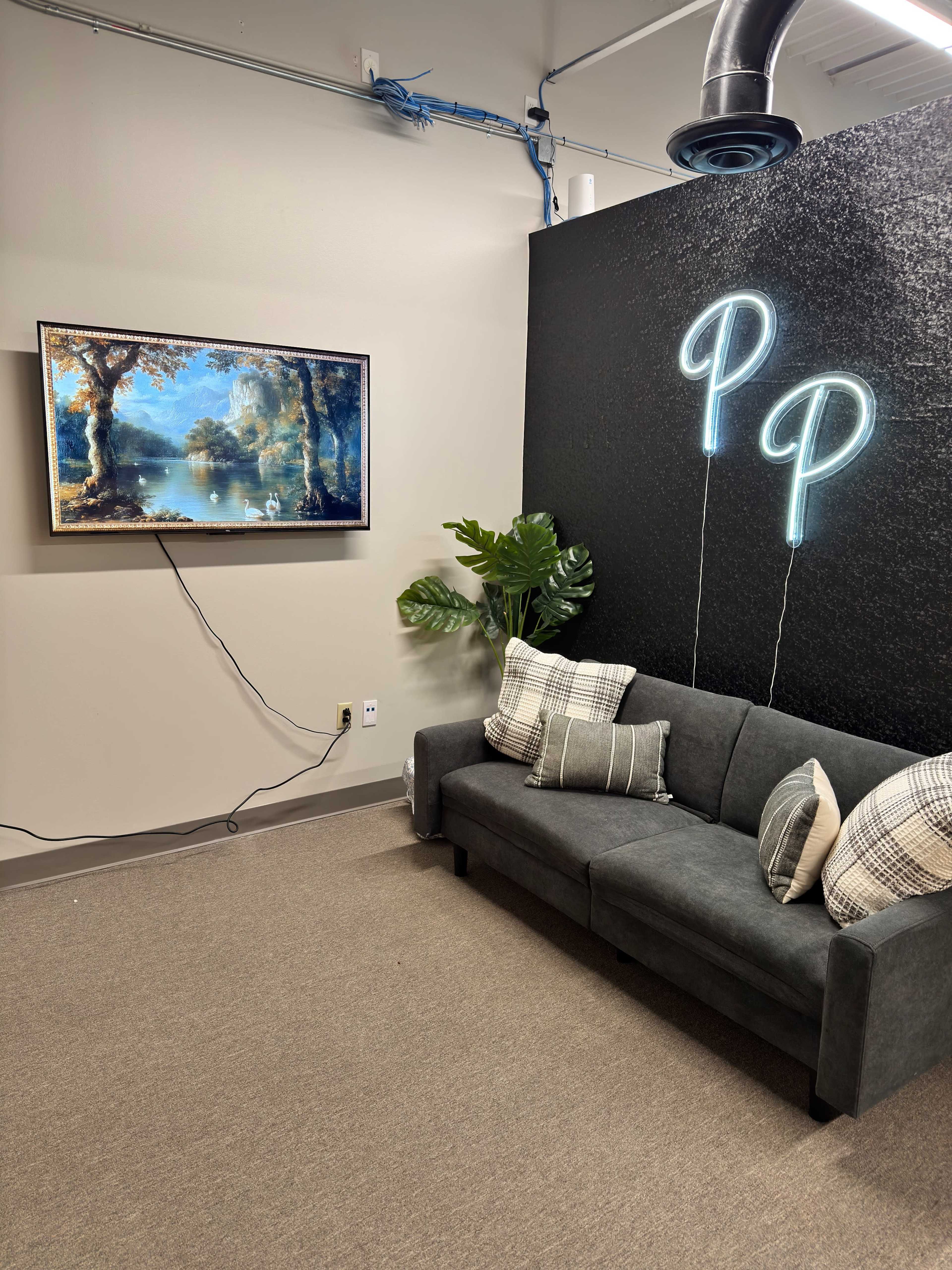 The image shows a cozy corner of a room featuring a dark-gray couch, a painting of a serene landscape on the wall, and two illuminated letters "P" on the wall beside a potted plant.