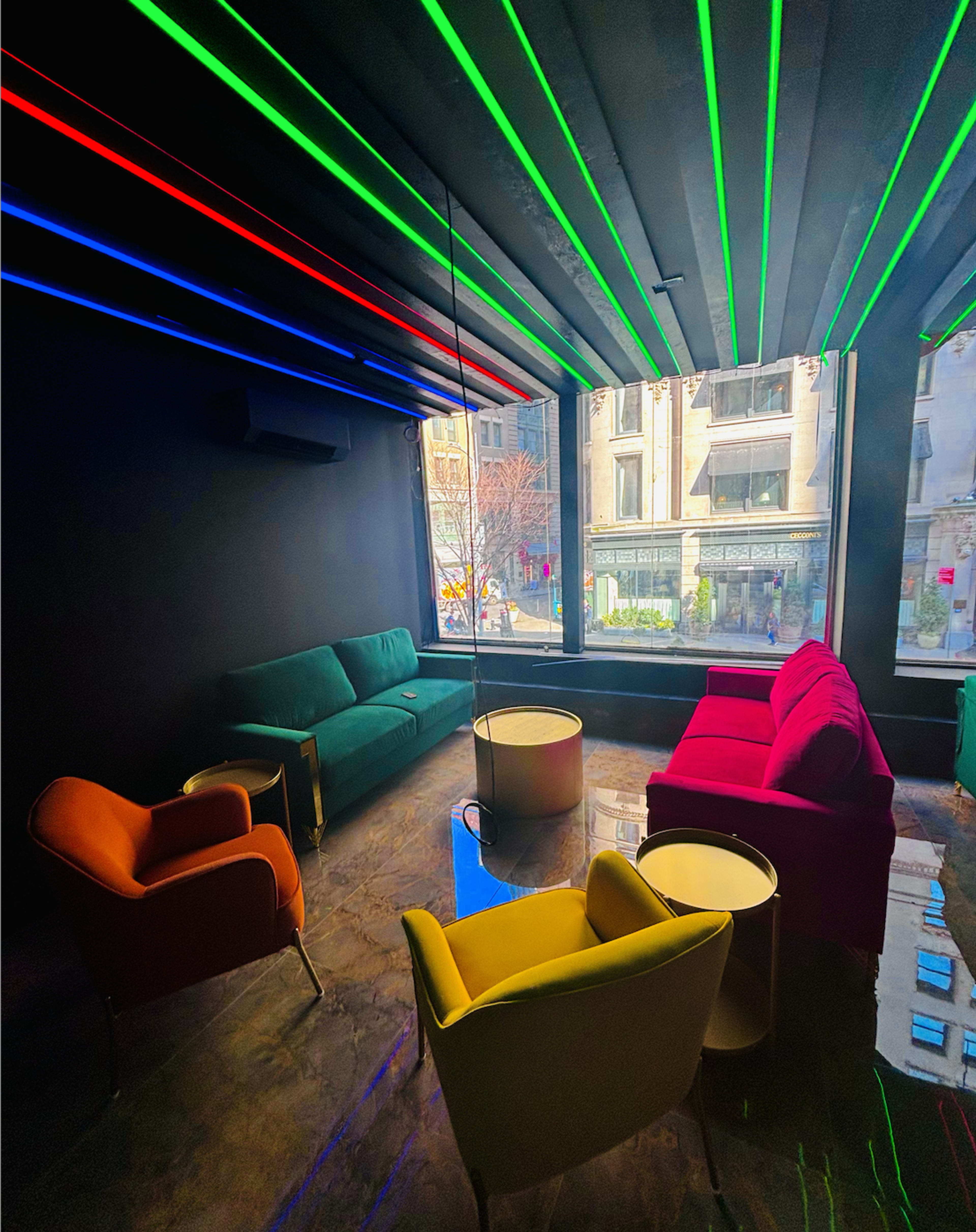 A modern lounge area with brightly colored furniture and neon lighting, overlooking a city street through large windows.