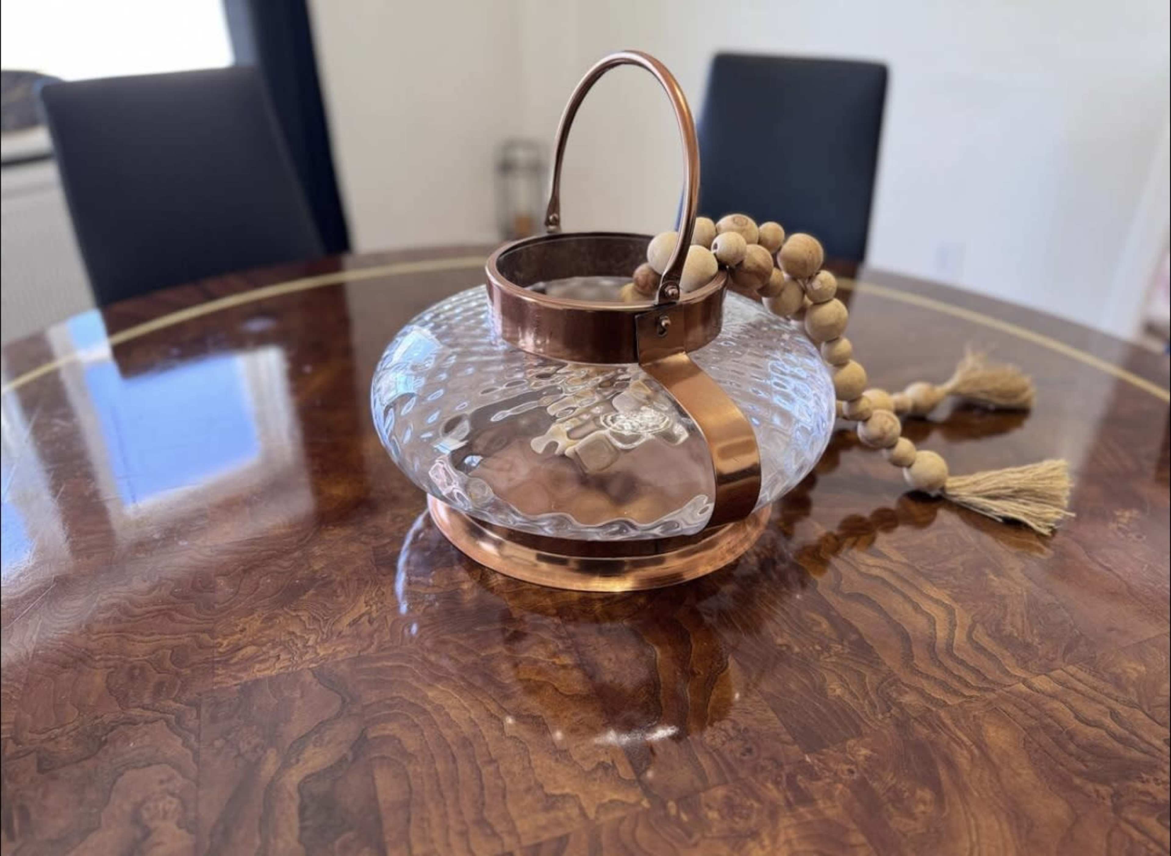 A decorative glass lantern with a copper handle and wooden beads sits on a round wooden table.