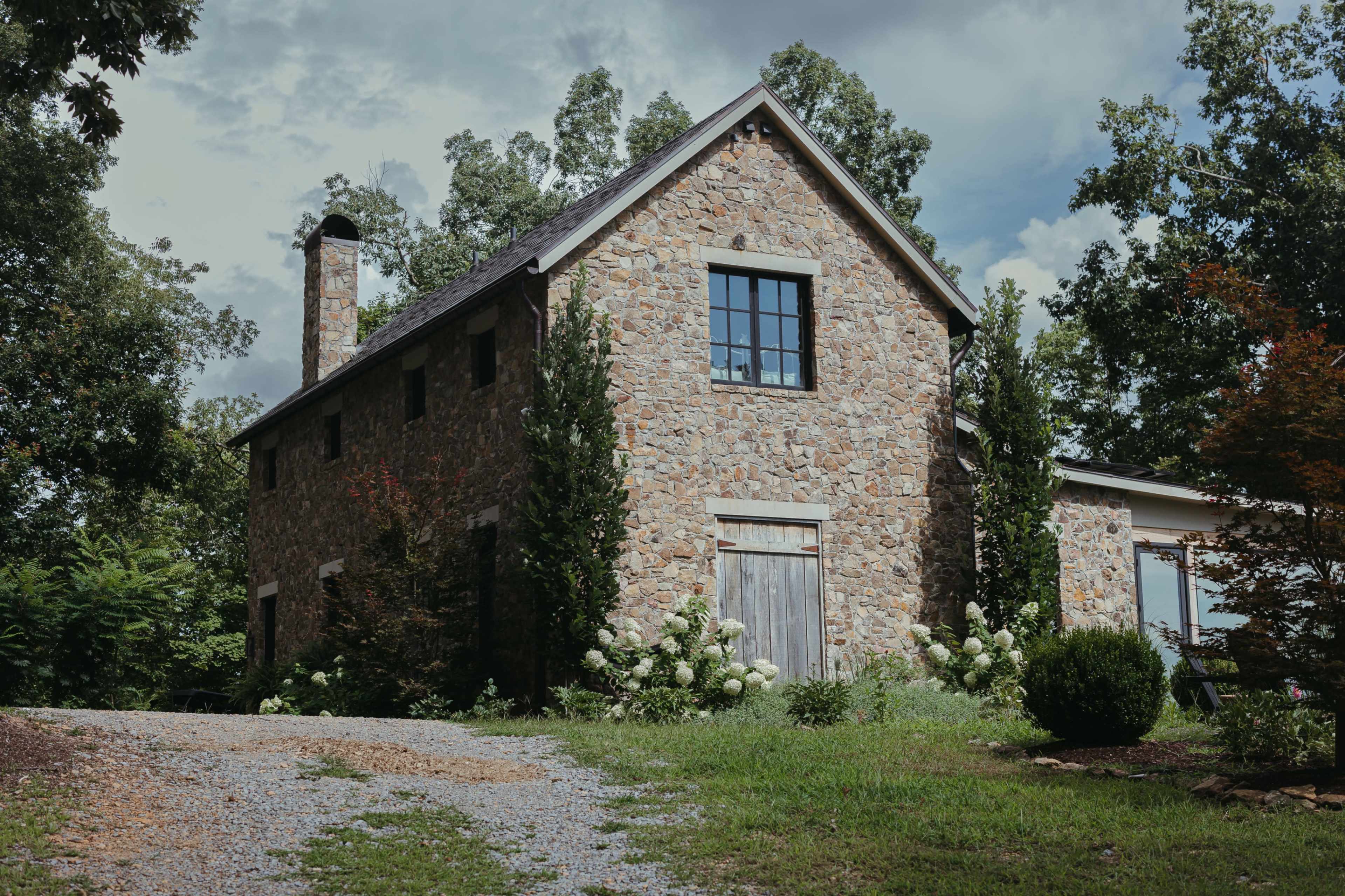 A stone cottage with a sloped roof and large windows is surrounded by greenery and a gravel path.