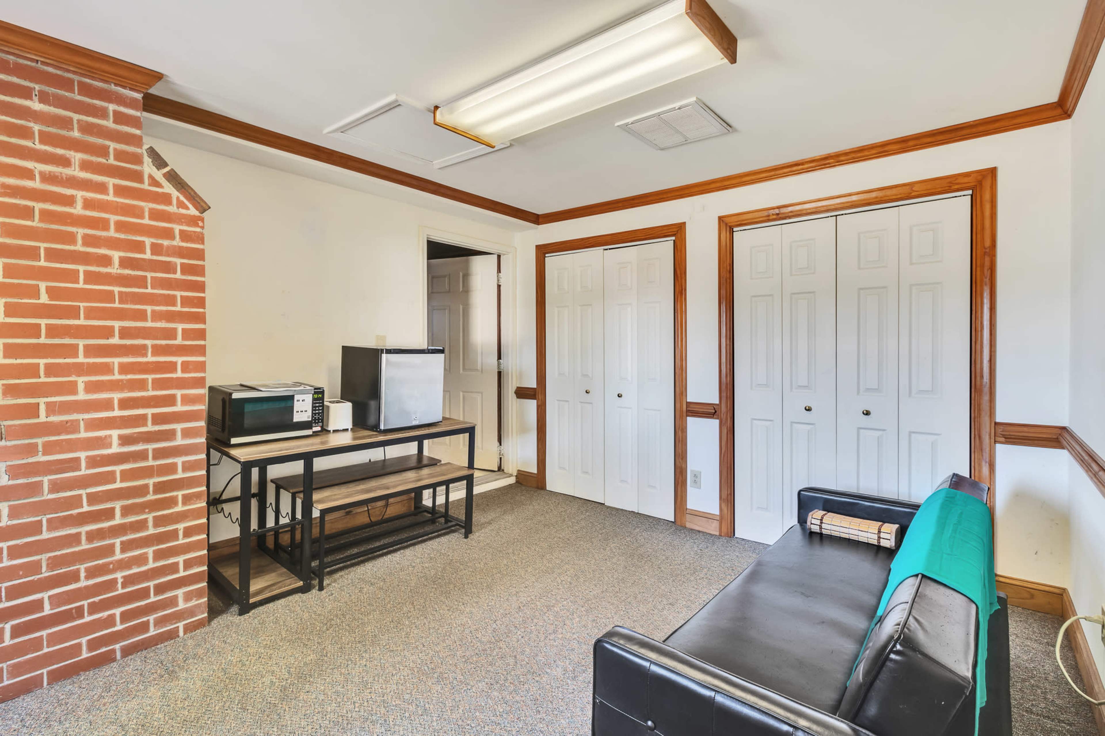 The image shows a small room with a brick wall, a black sofa, a table with a microwave and a TV, and two closed closet doors.