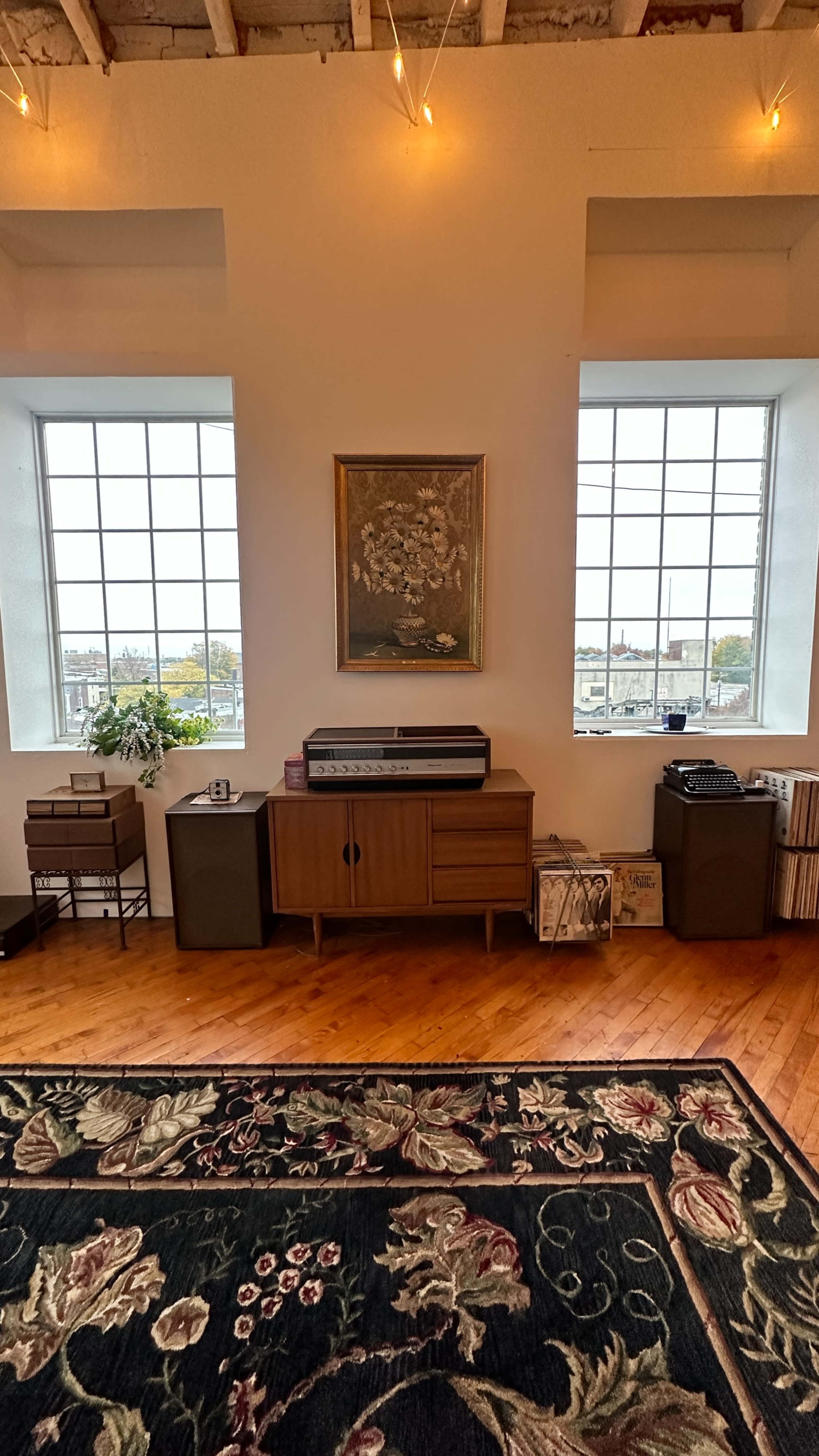 A room with wooden flooring, two large windows, a vintage stereo cabinet, and decorative elements including a painting and a rug.