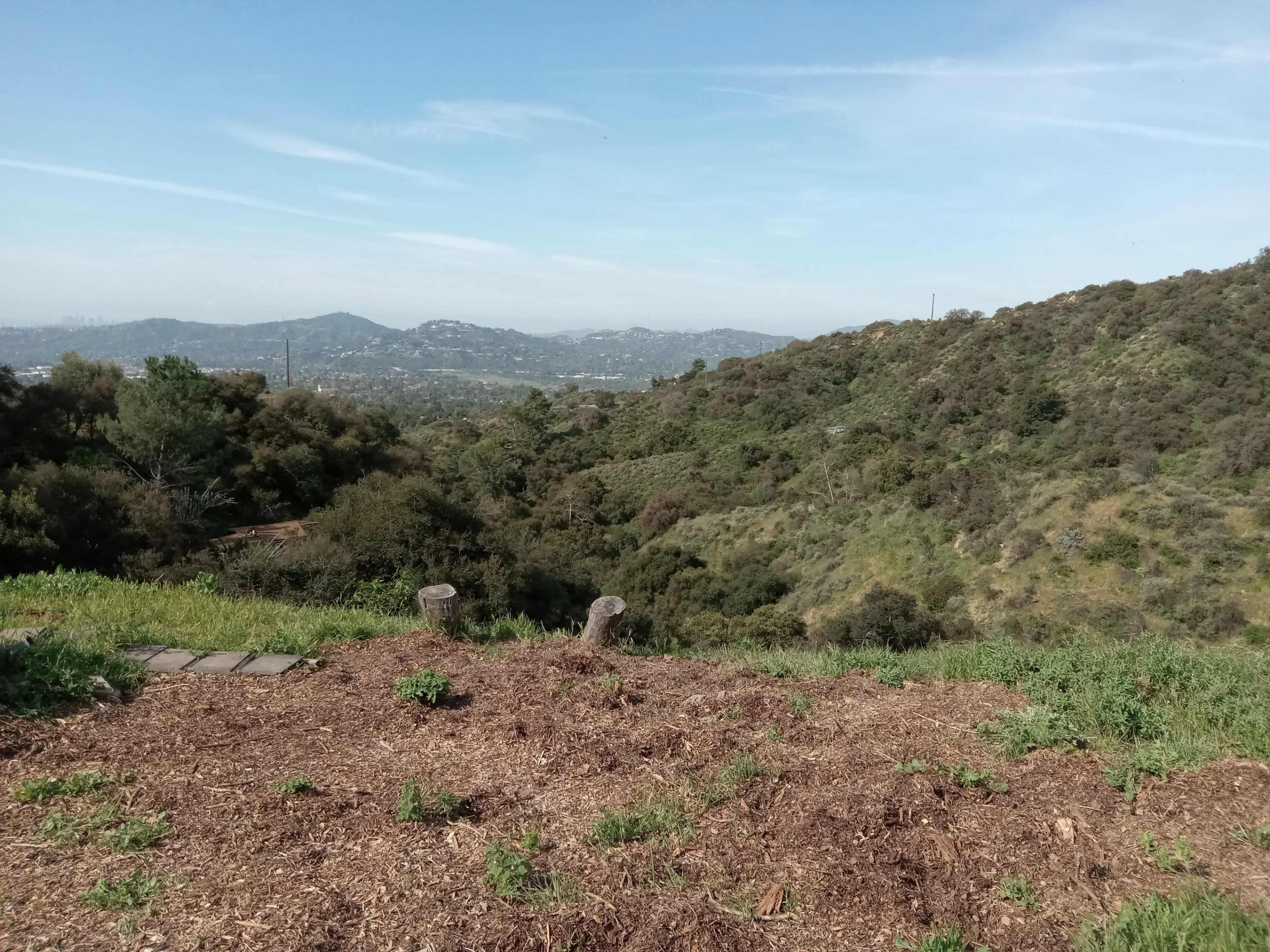 The image shows a hillside with green vegetation and a view of distant mountains and hills under a clear sky.