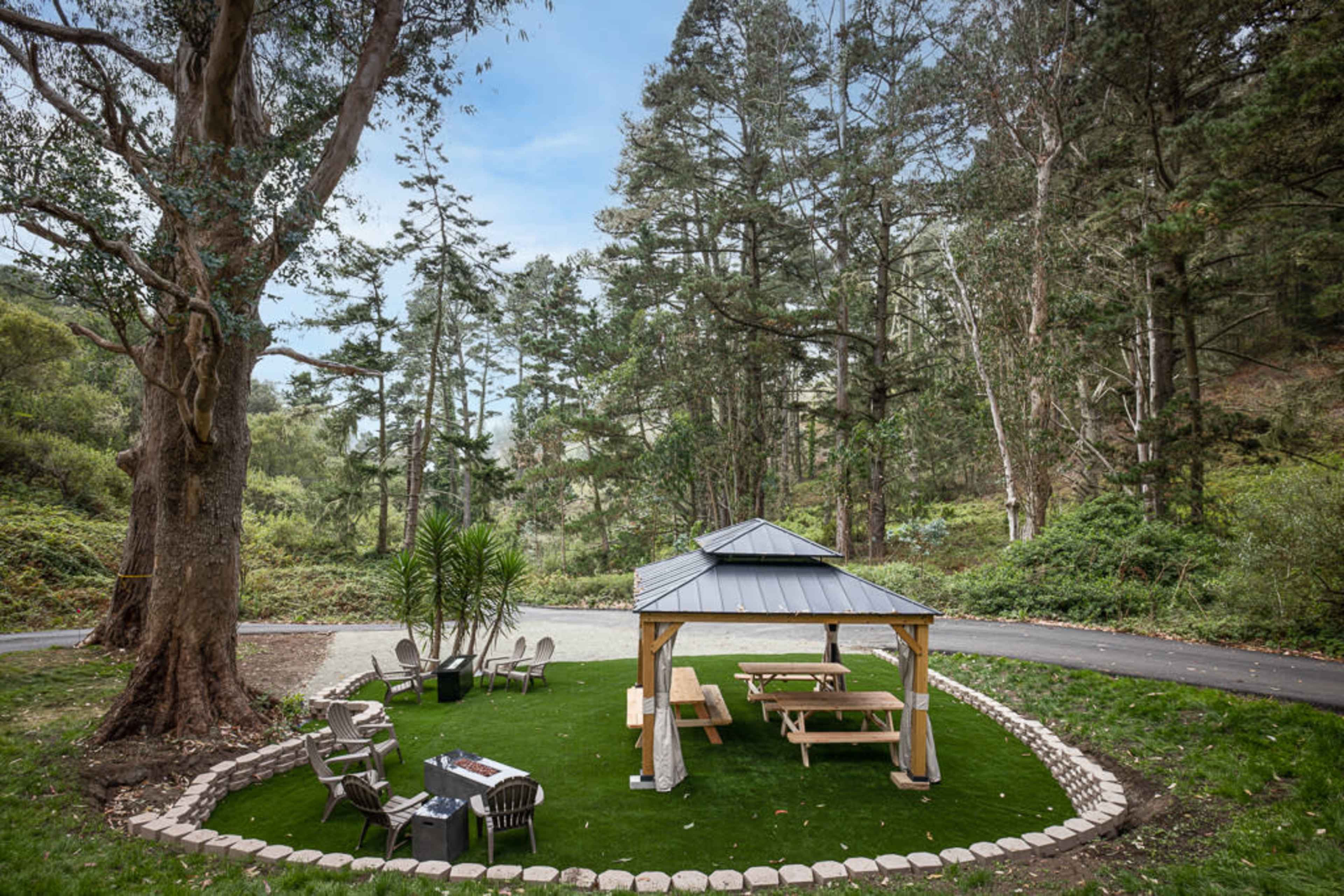 A gazebo with picnic tables, surrounded by trees and grass, in a cleared area of a forest.