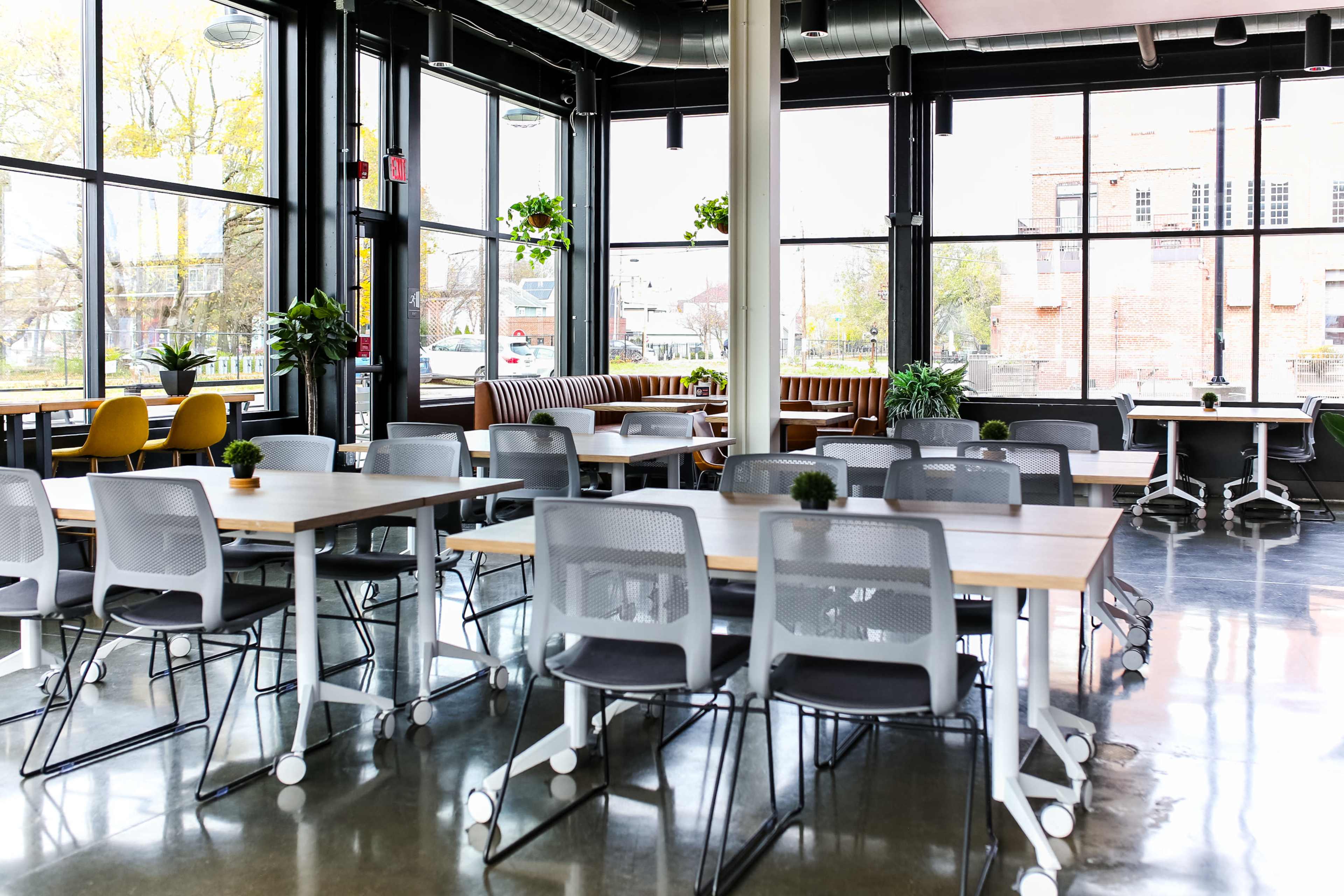 The image shows a modern office space with multiple tables and chairs arranged in a bright room filled with natural light from large windows.
