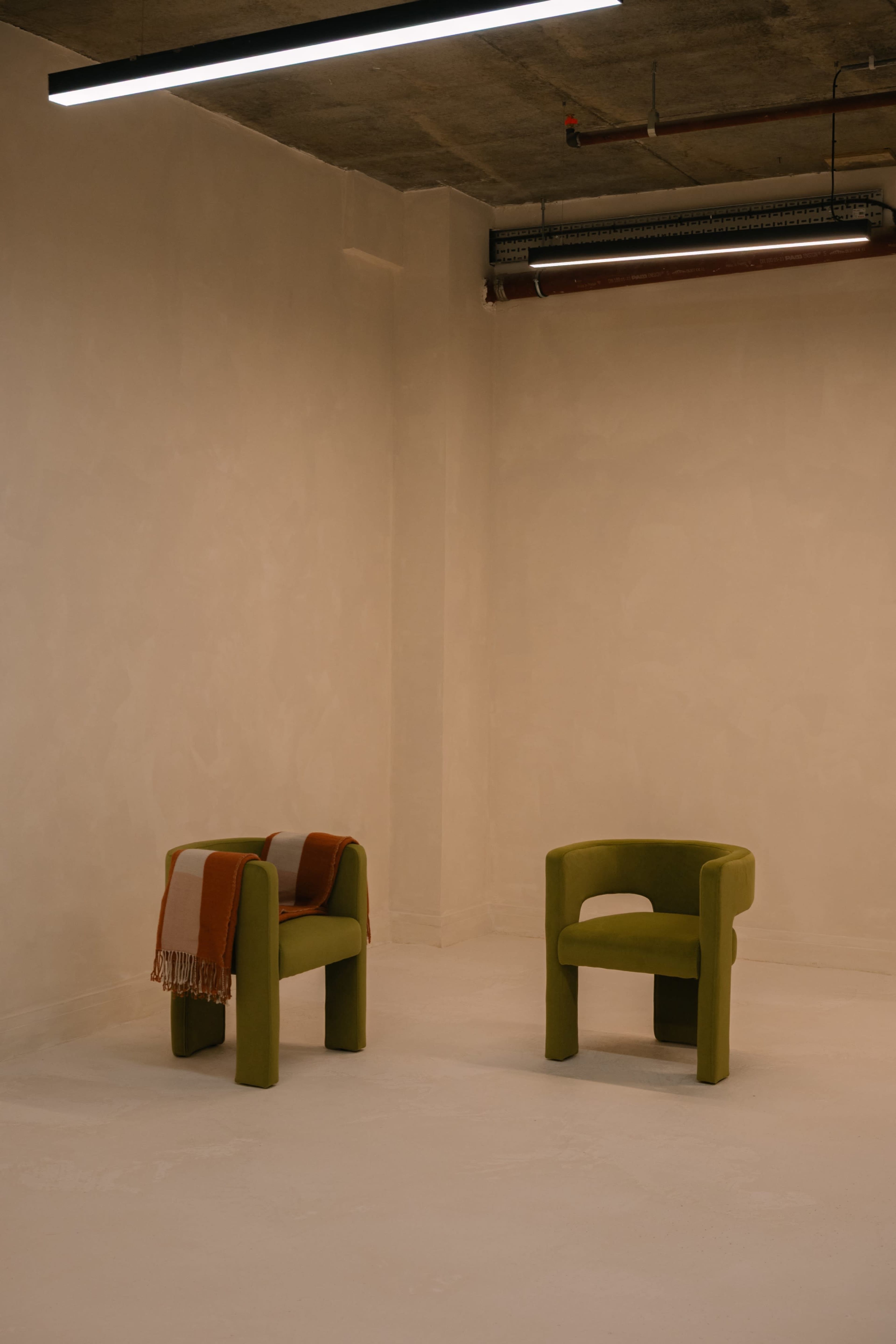 Two modern chairs, one green and one with a blanket draped over it, are positioned against a plain beige wall in a minimalist room.