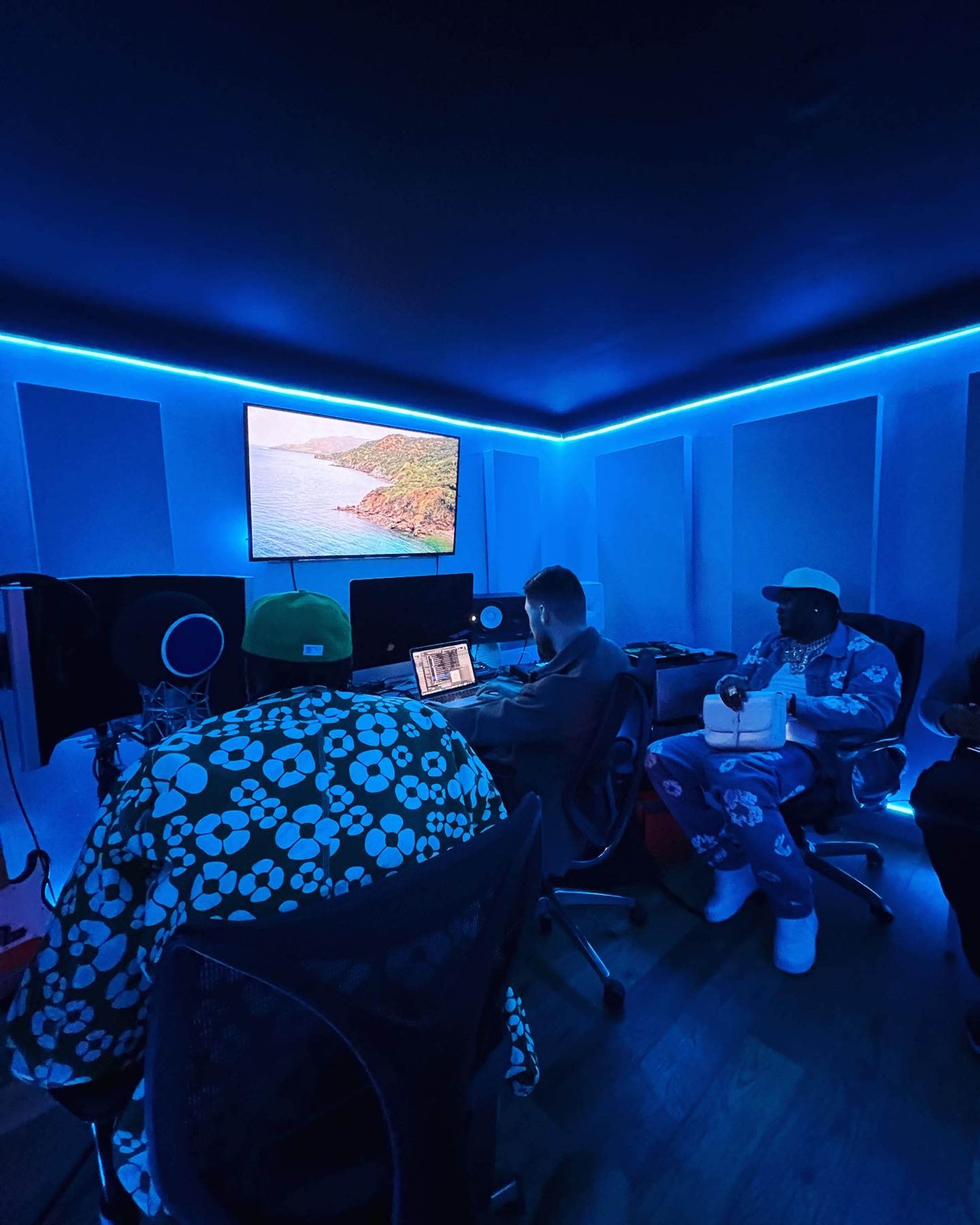 The image shows a recording studio with three individuals focused on their laptops, surrounded by blue LED lighting and a large screen displaying a scenic landscape.