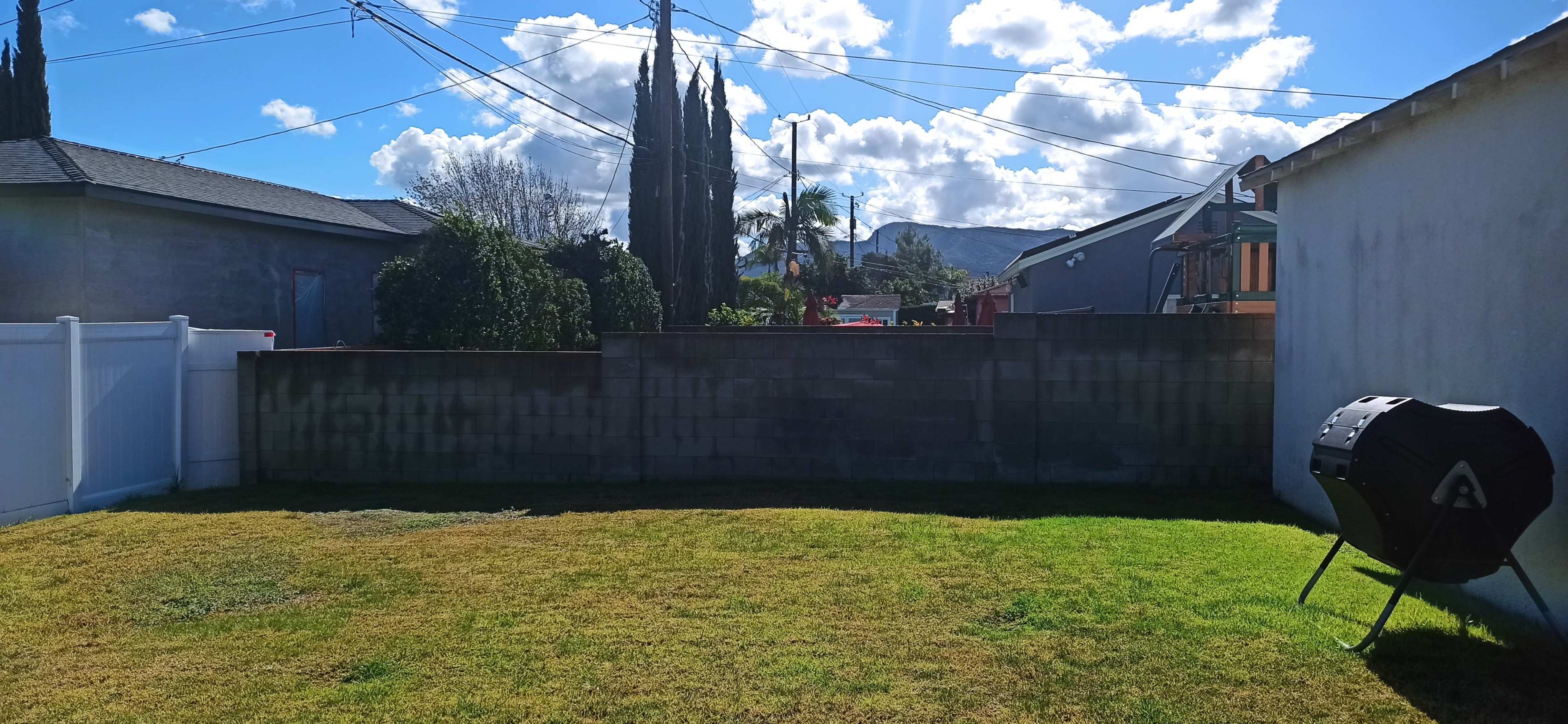The image shows a green backyard with a low stone wall, a black grill on a stand, and a clear blue sky with scattered clouds.