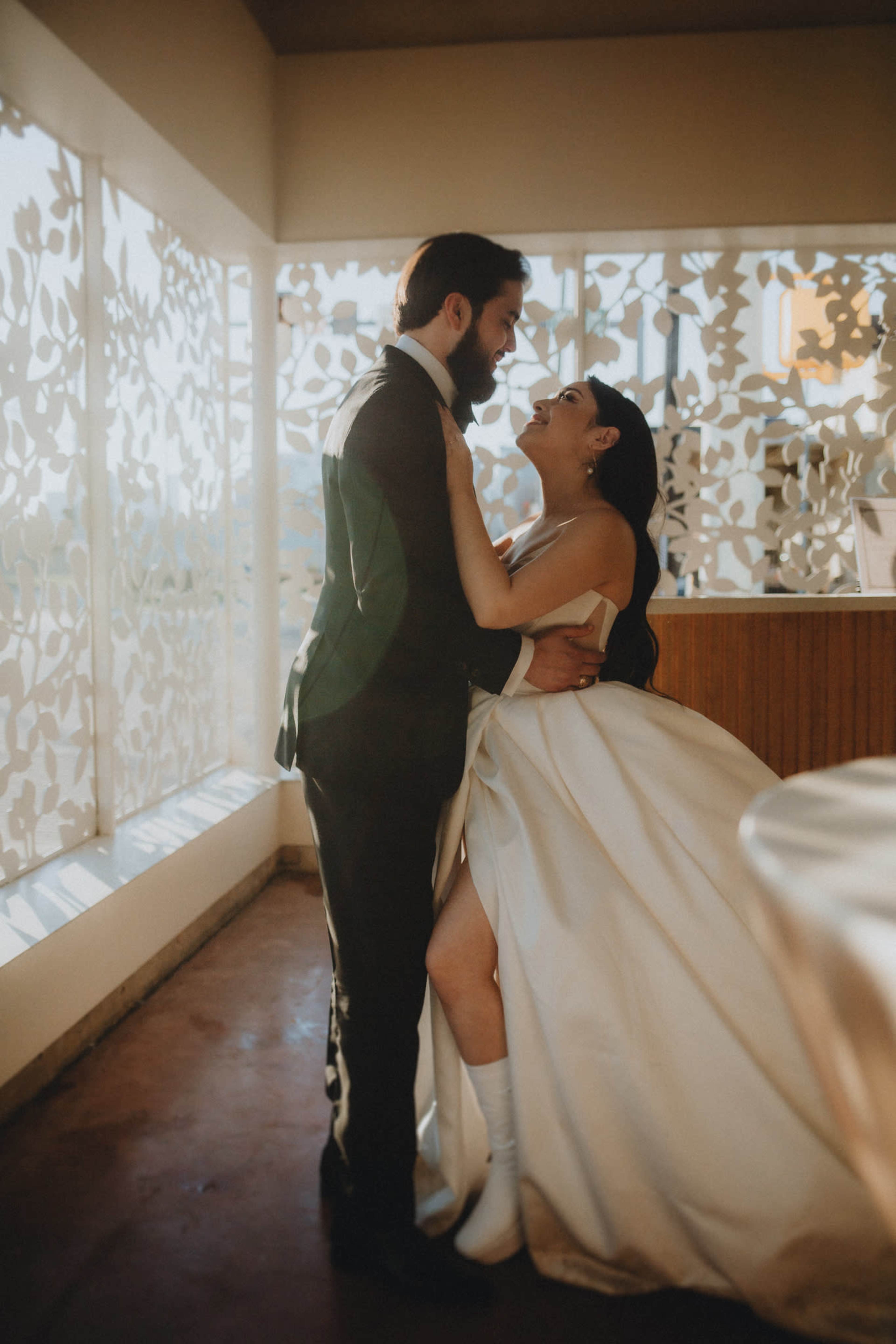 A bride and groom embrace warmly in a brightly lit room with decorative foliage-patterned windows.