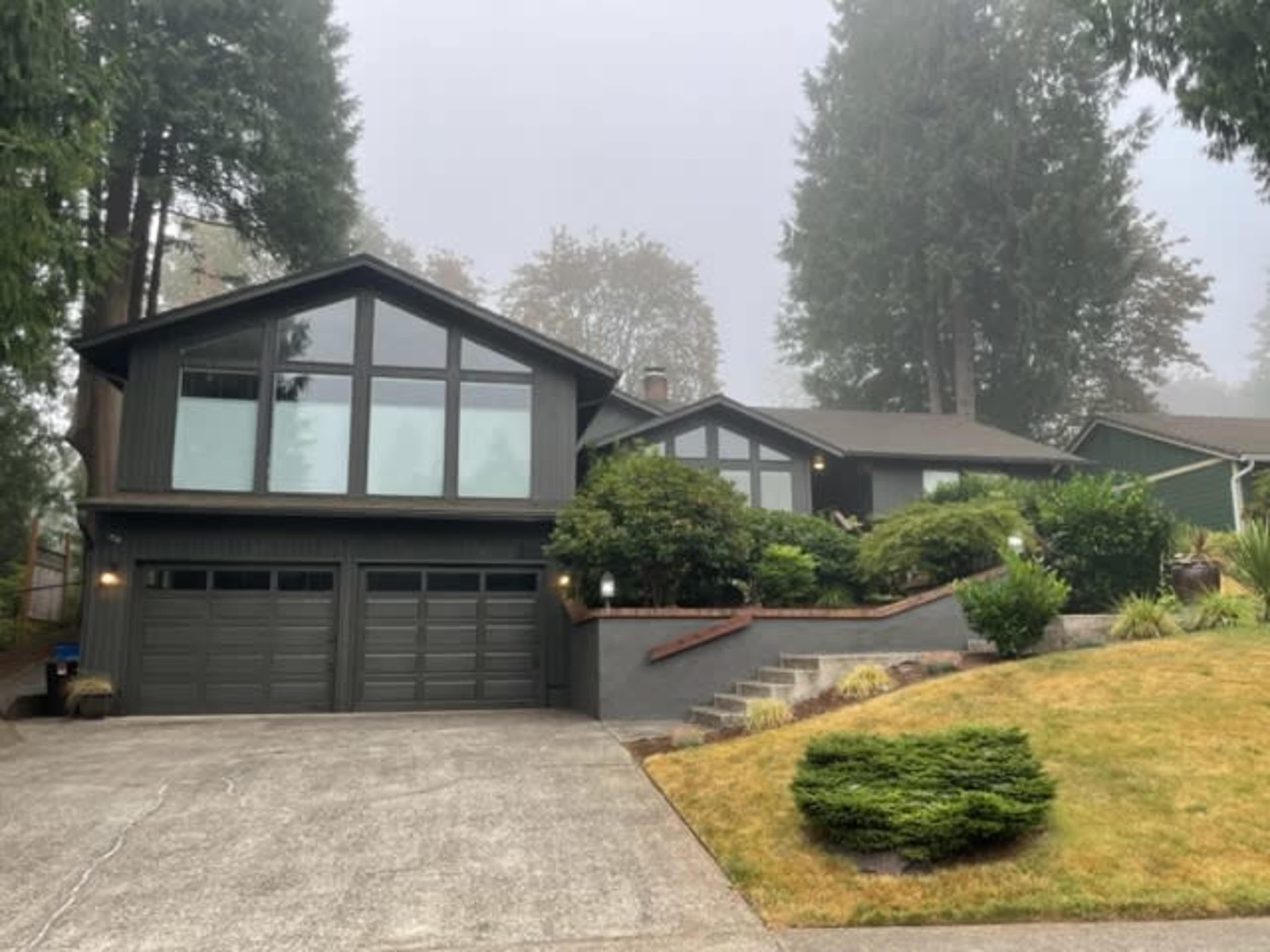 A modern two-story house with large windows and a two-car garage is set on a landscaped lot surrounded by tall trees in a foggy atmosphere.