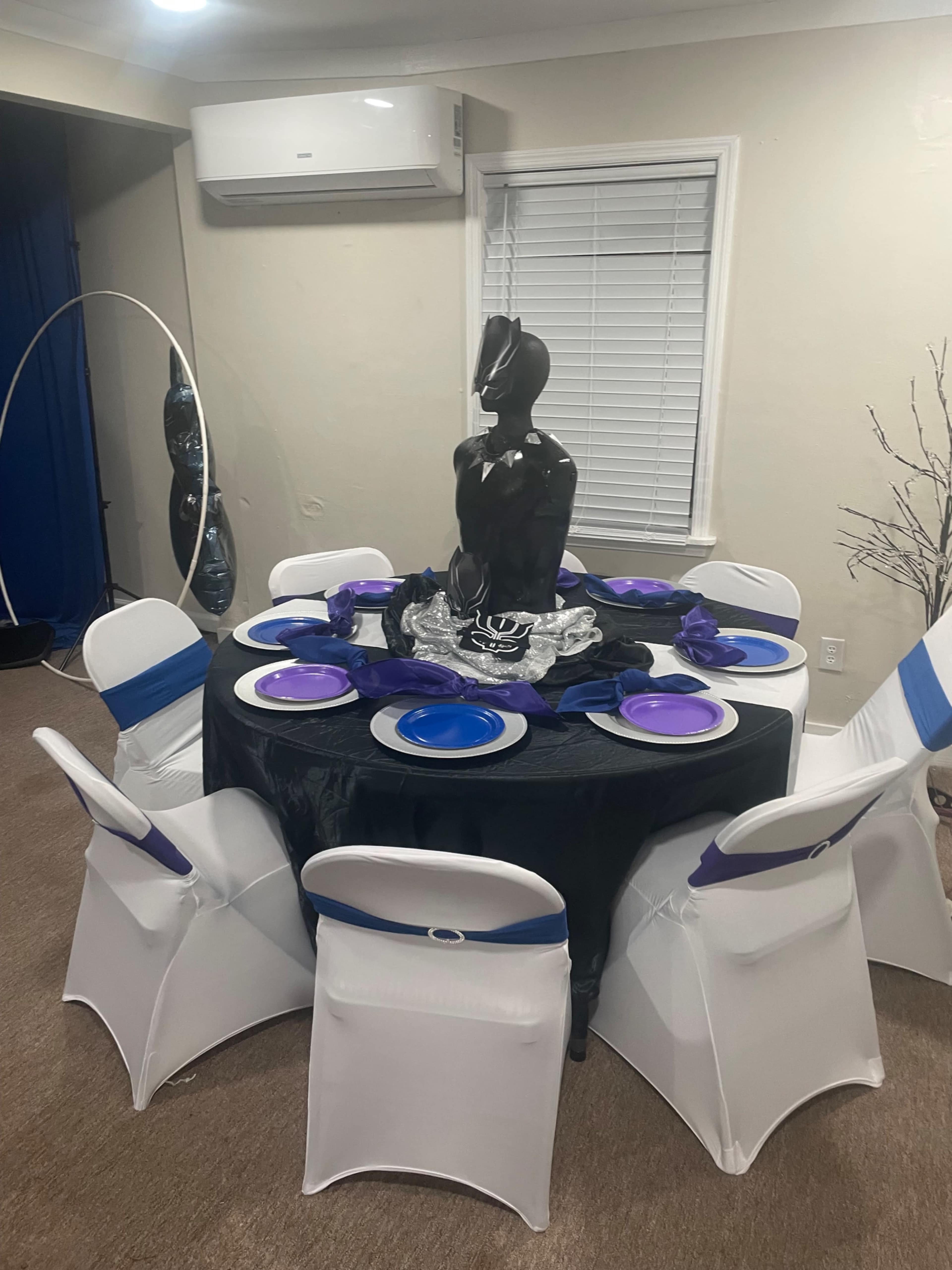 A round table with black and purple decorations is set with plates and a central black statue surrounded by six chairs.