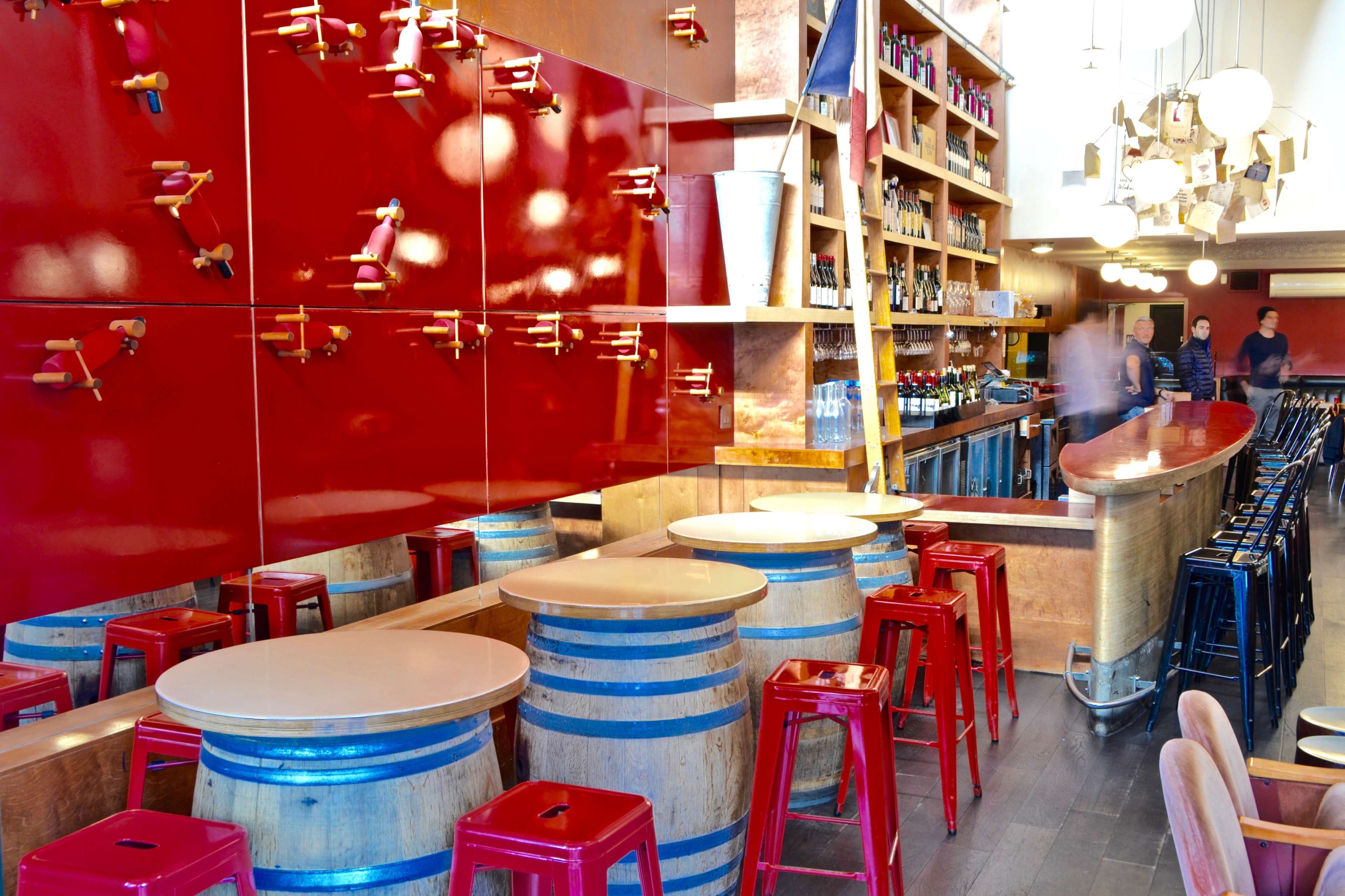 A modern bar with red walls, wooden barrels as tables, and sleek seating arrangements.