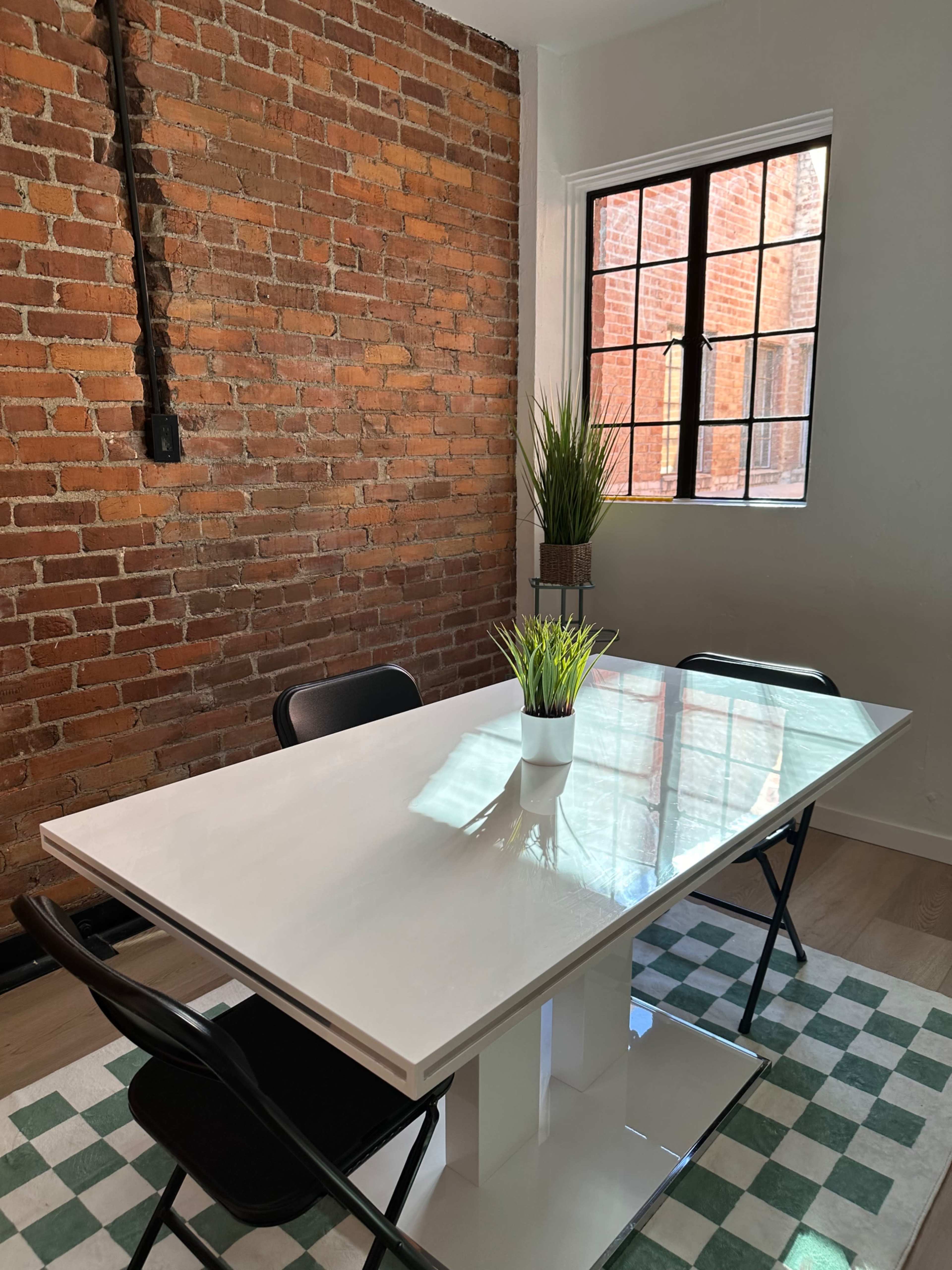 A modern white table with two black chairs is positioned on a green and white checkered rug against a backdrop of exposed brick walls and a window.