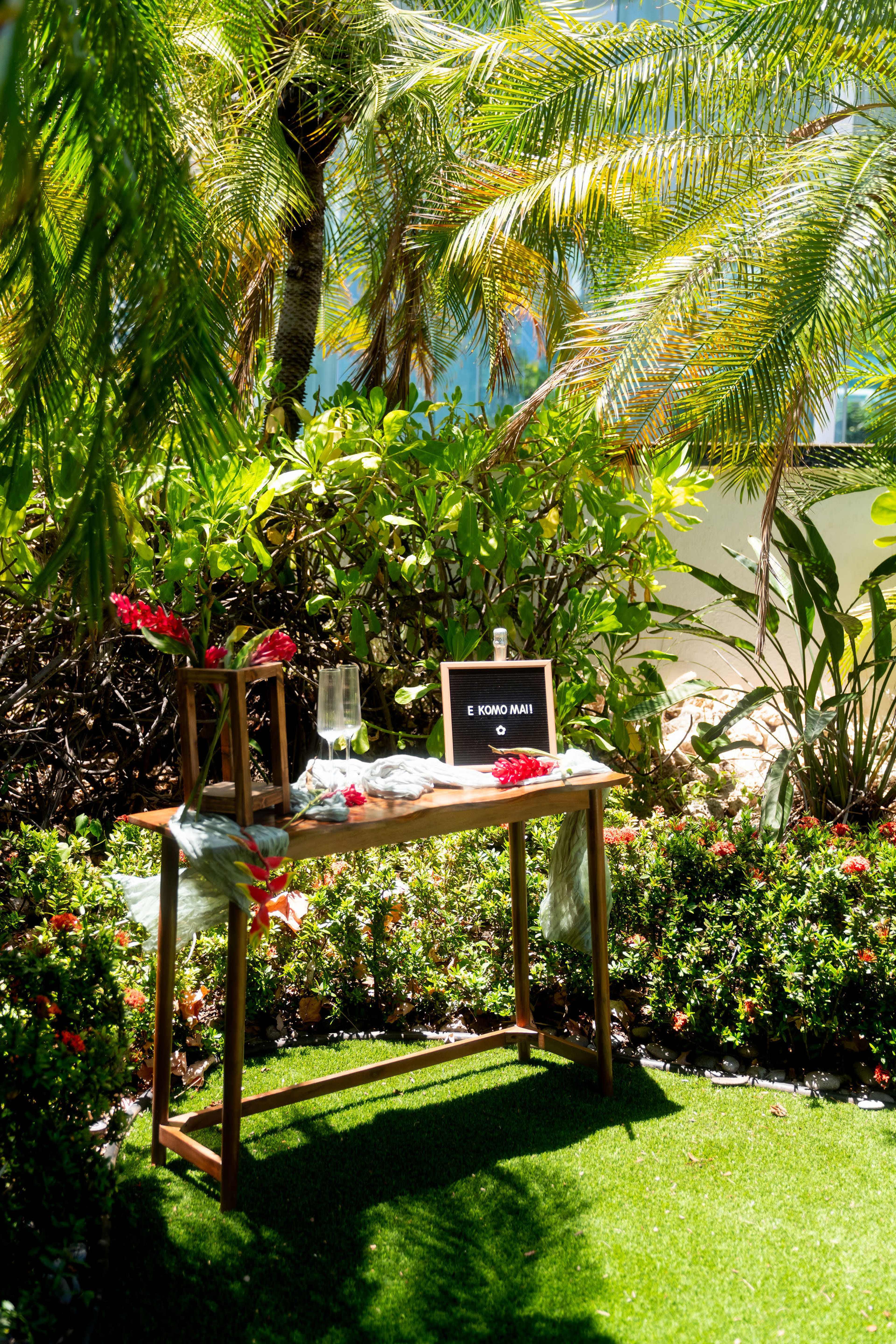 A wooden table is set outdoors among lush greenery, featuring a small sign that reads "E KOMO MAI" and decorated with flowers and a glass.