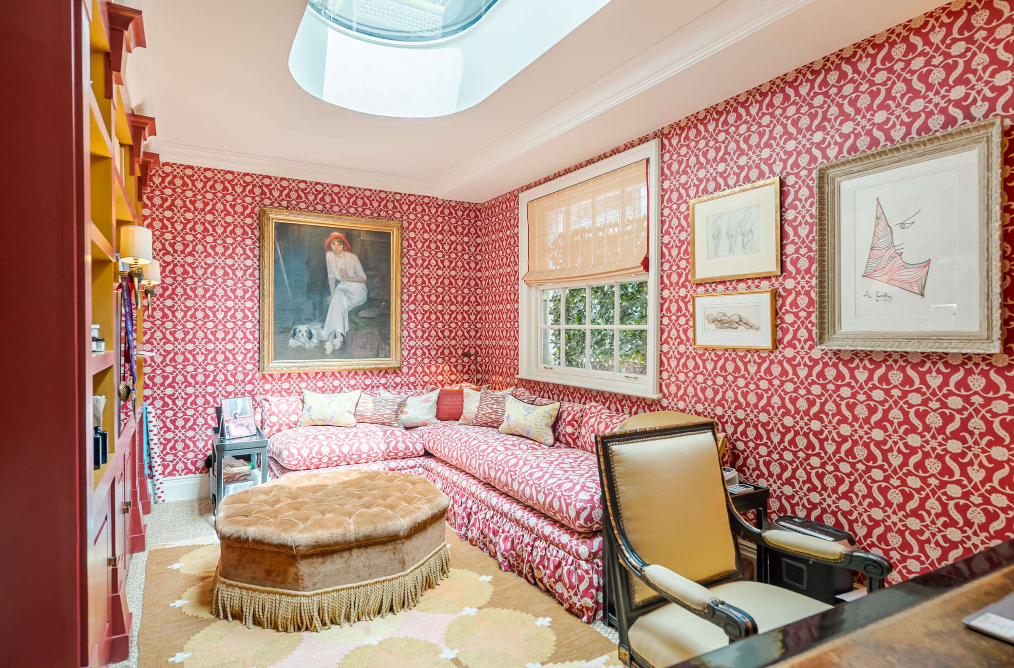 The image shows a cozy room with patterned red wallpaper, a sectional sofa, and a skylight, featuring artwork on the walls and a round ottoman in the center.
