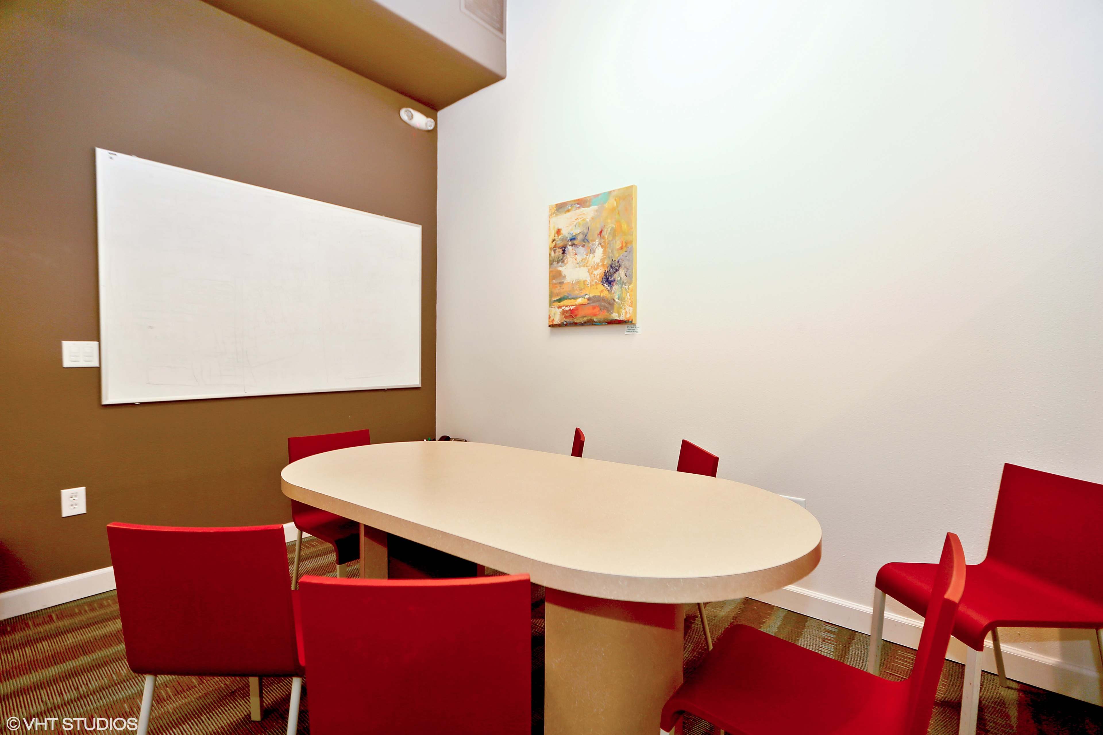 A small conference room features a large oval table surrounded by red chairs and a whiteboard on one wall.