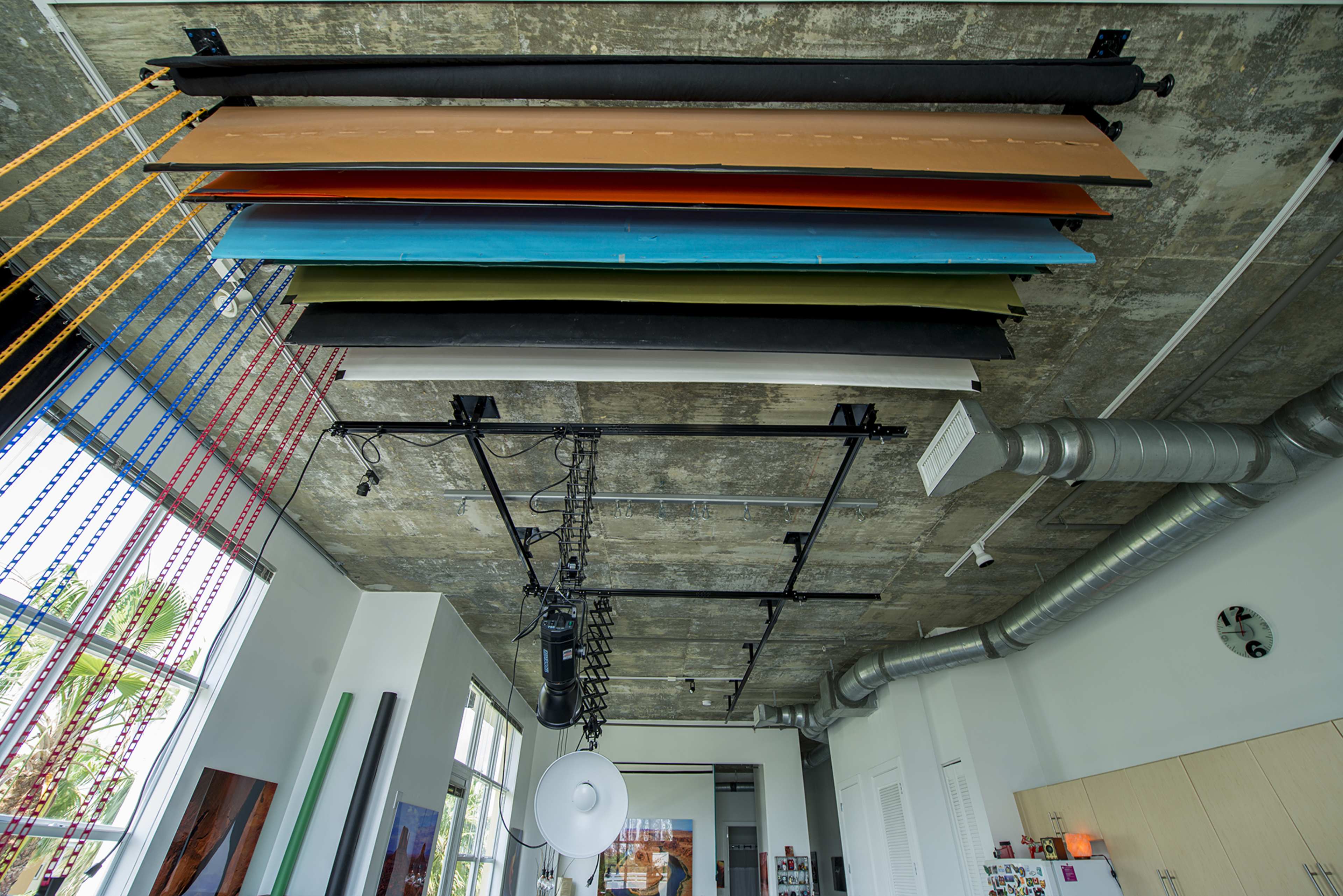 The image shows a ceiling with colorful sheets suspended overhead in an industrial-style room featuring exposed pipes and large windows.