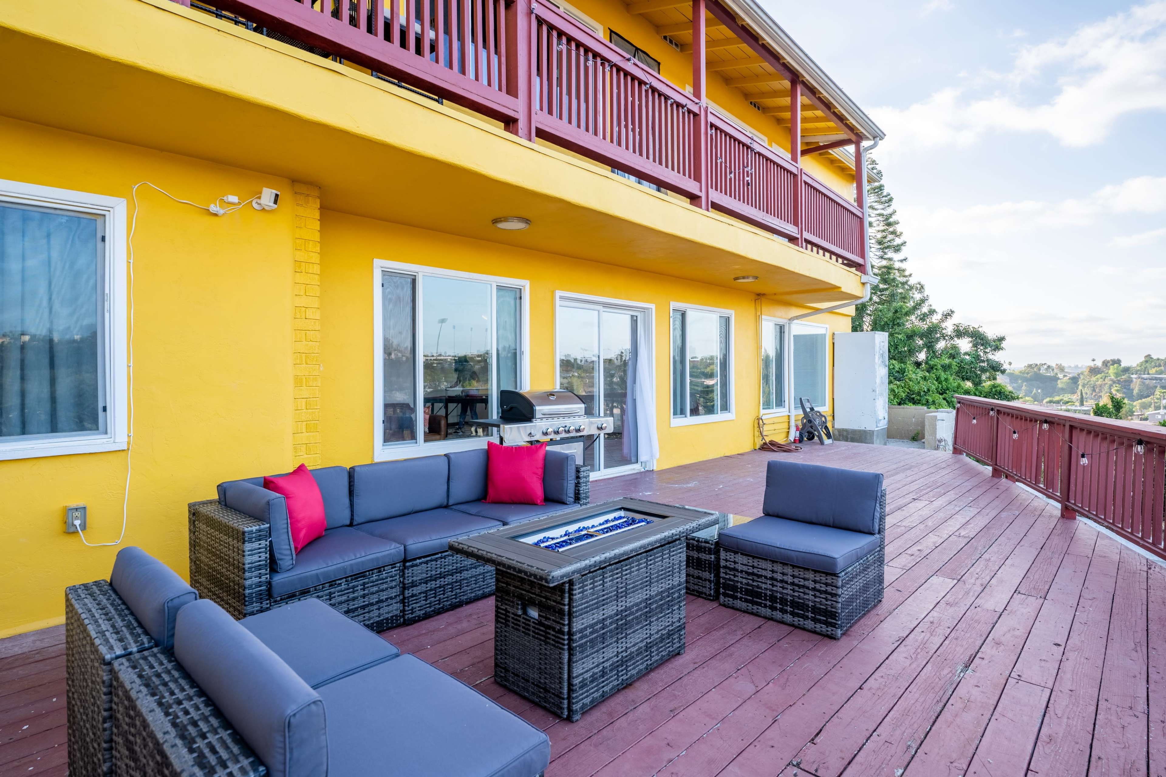 The image shows a deck with a seating area consisting of a dark wicker sofa set and a fire pit, adjacent to a yellow building with a second floor balcony.