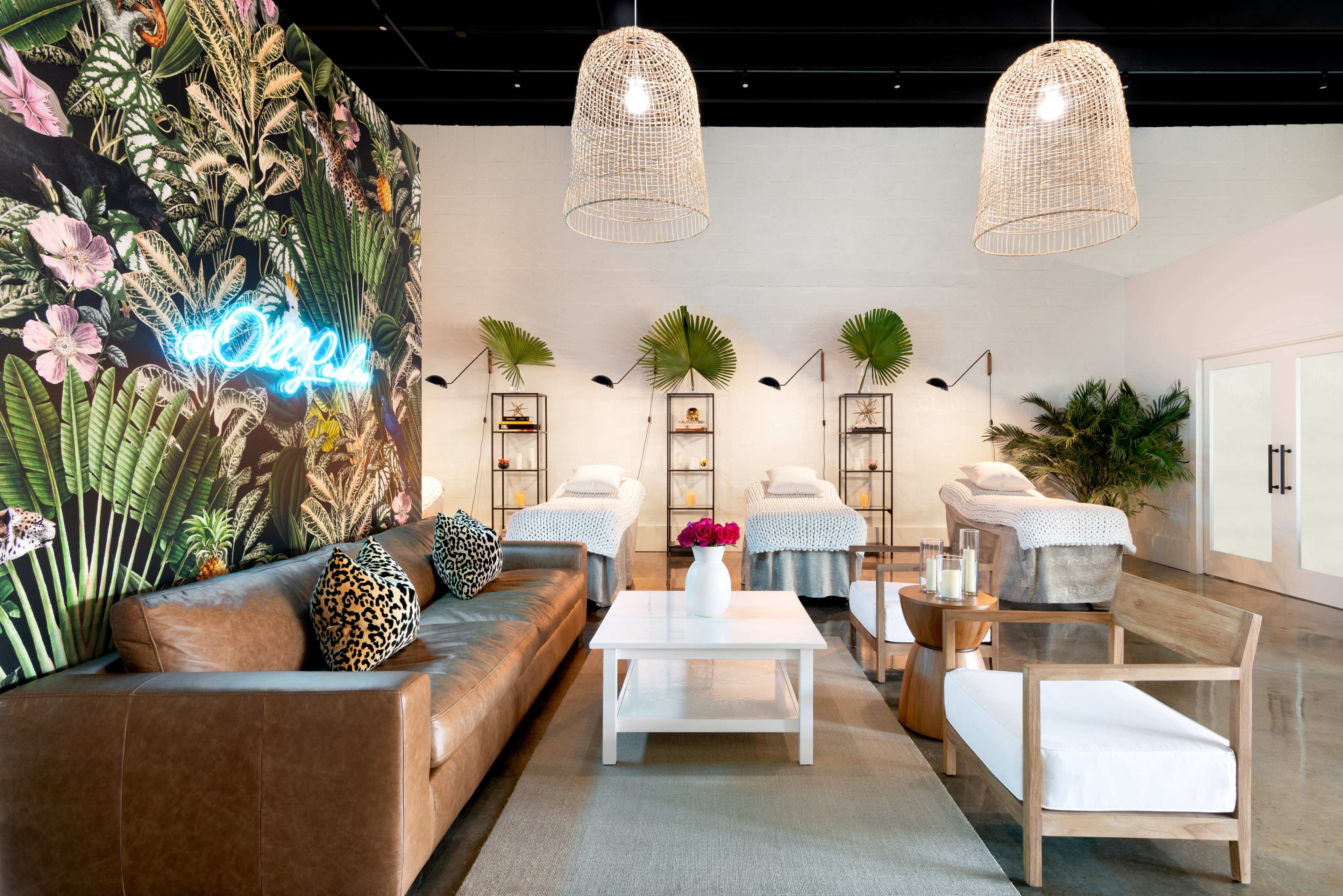 A modern spa interior featuring a leafy accent wall, several treatment beds arranged in a line, and stylish seating areas with pendant light fixtures overhead.