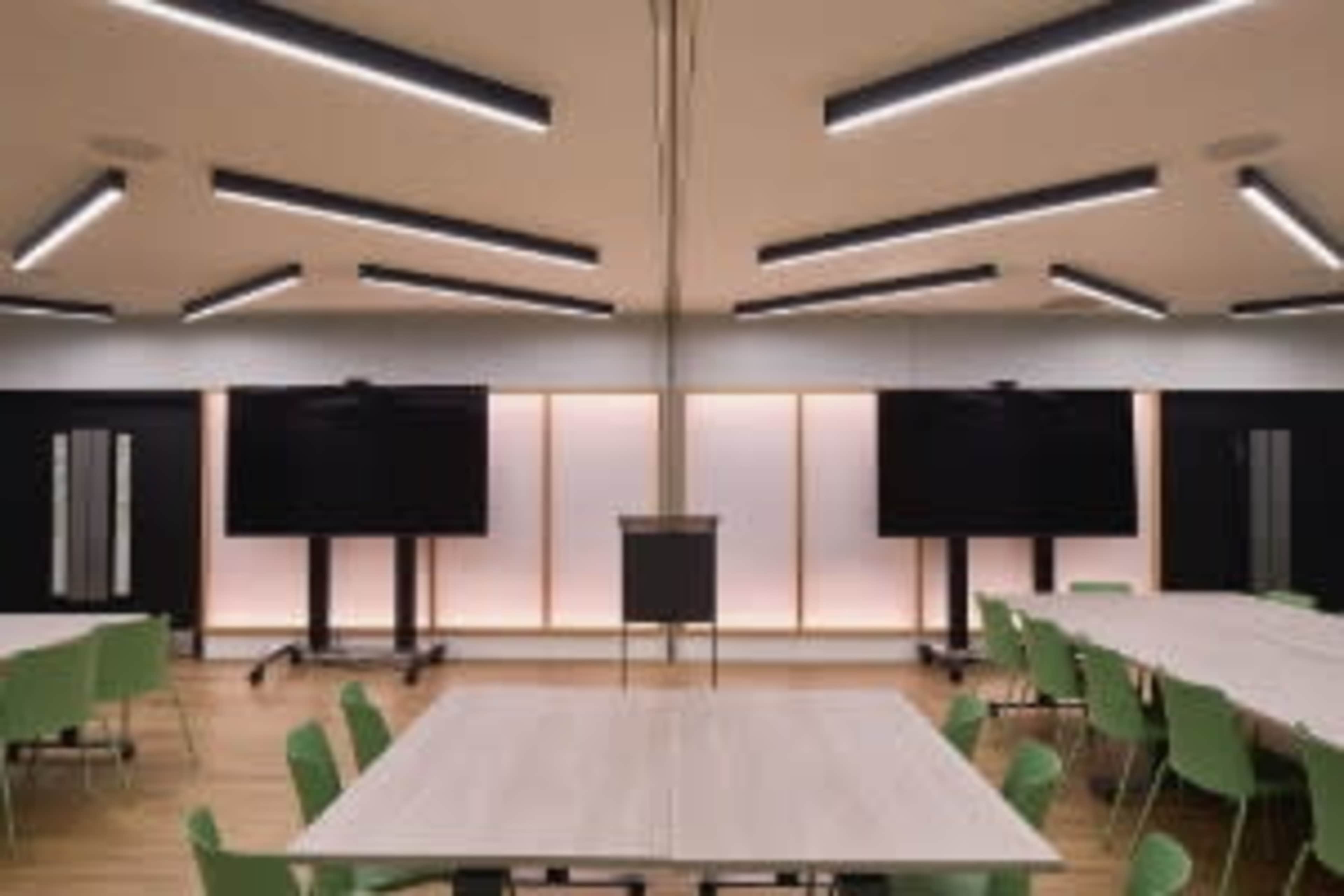 The image shows a modern conference room with several tables and green chairs arranged around them, along with two large screens on the walls.