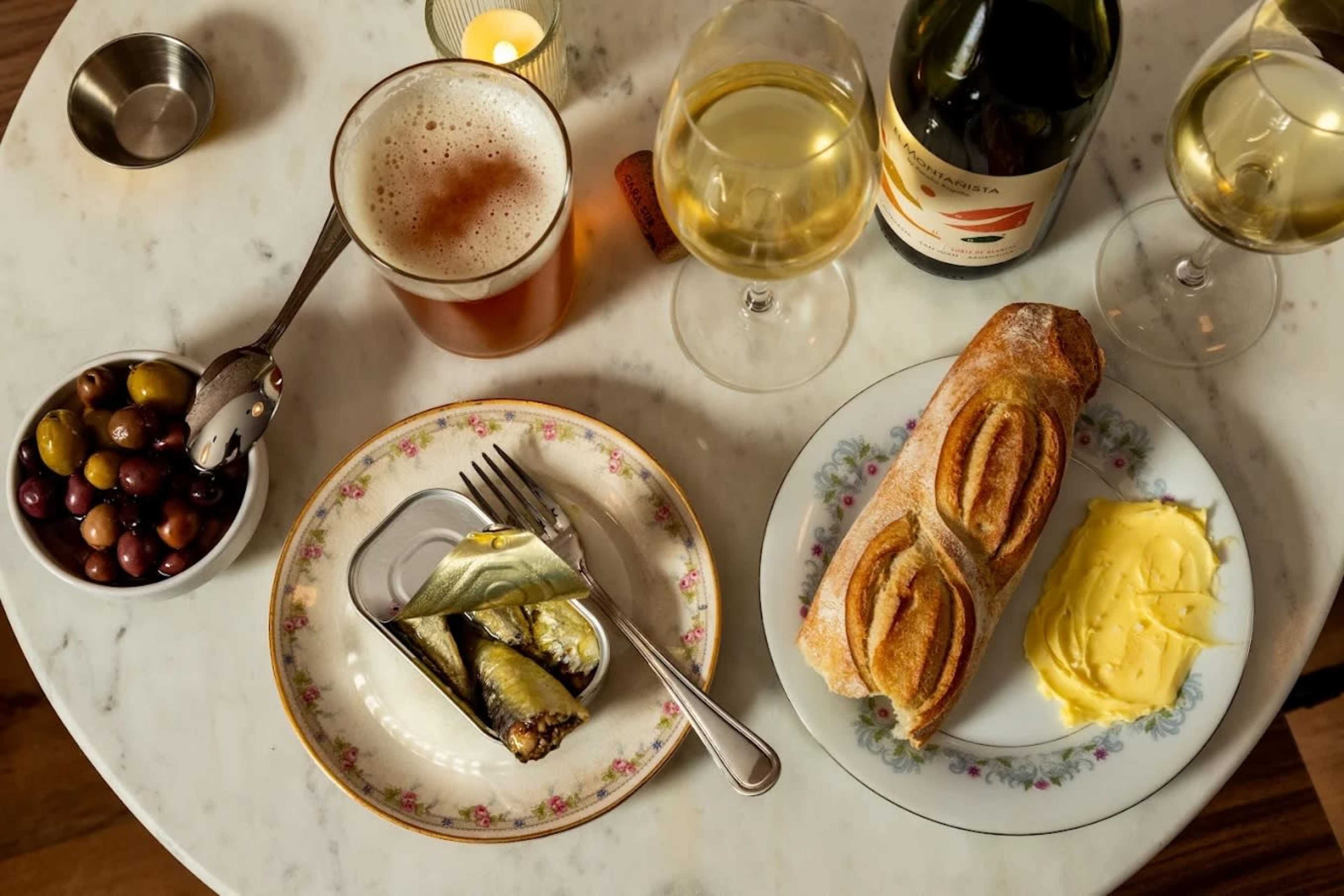 The image shows a marble table with a plate of olives, a dish of sardines, a roll with butter, and two glasses of white wine alongside a glass of beer.