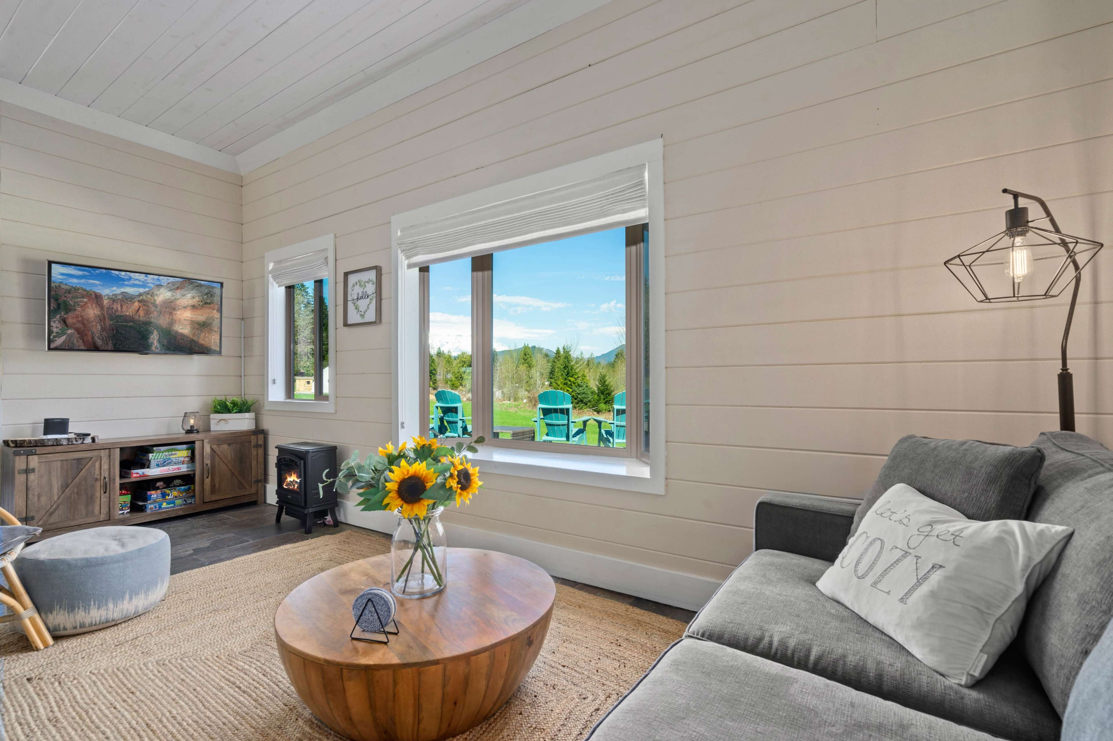 A cozy living room features a gray sofa, a wooden coffee table, and large windows overlooking a patio with green chairs.