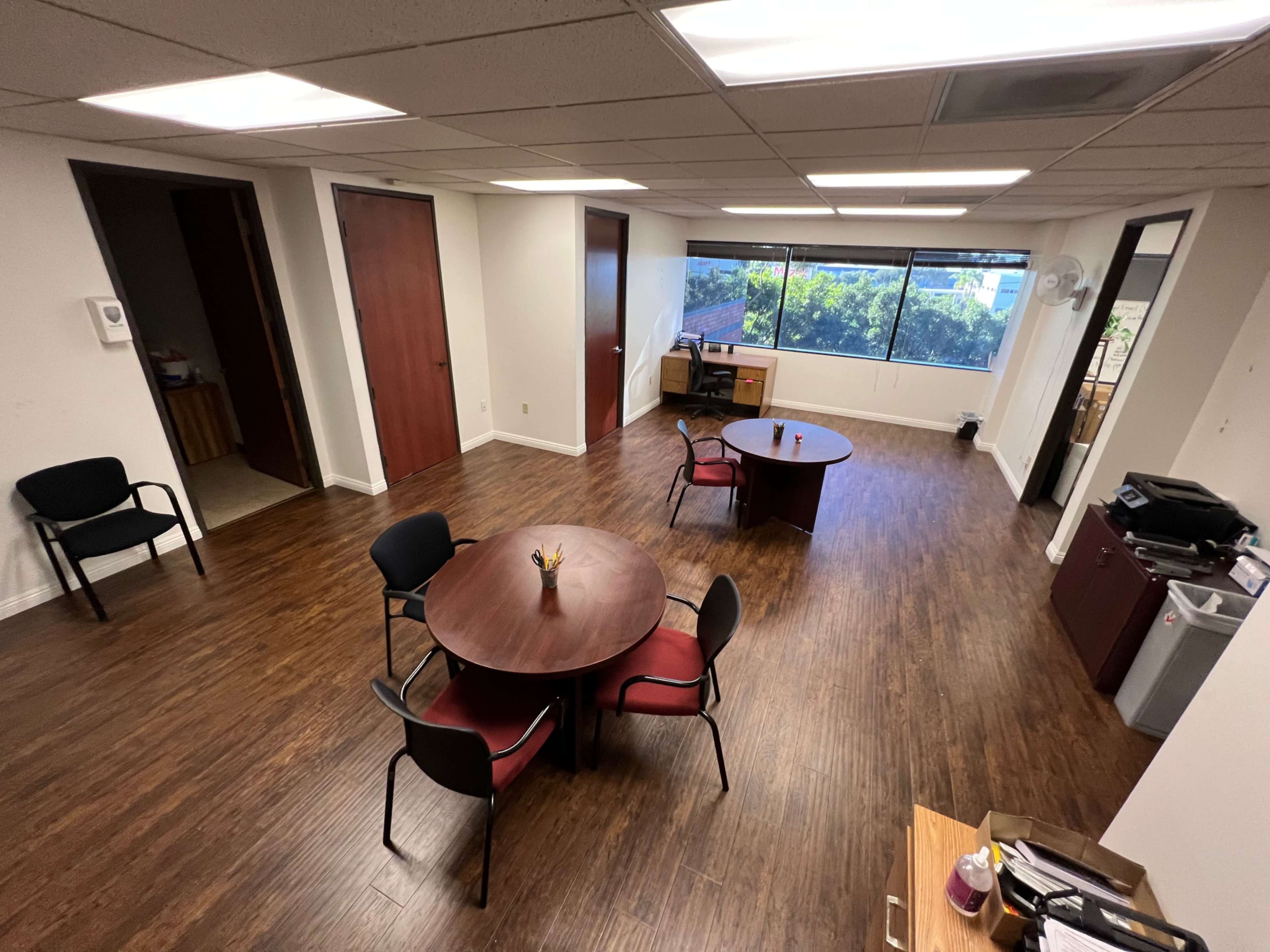 An office space featuring two round tables with chairs, a desk near a window, and wooden flooring.