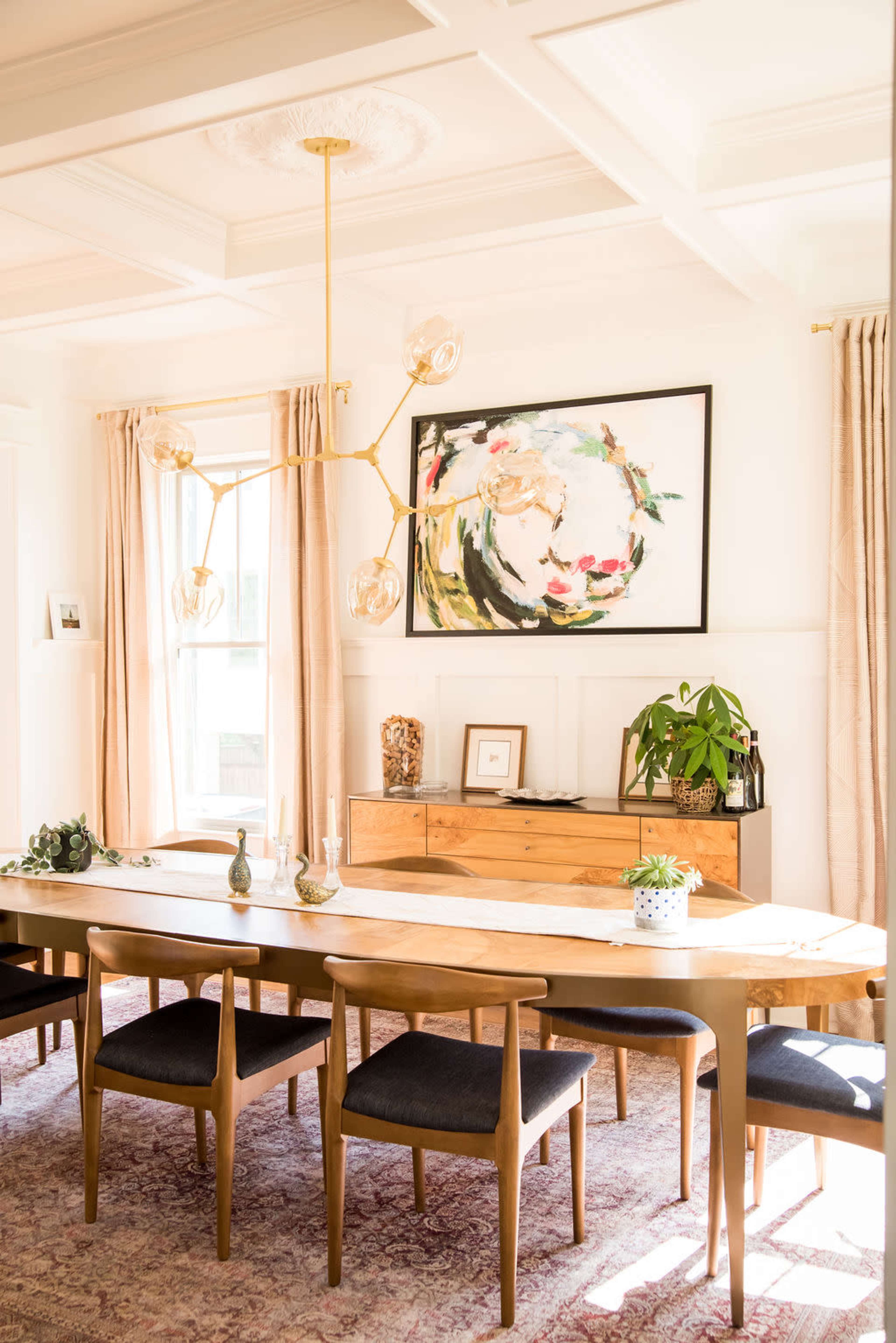 A bright dining room features a long table surrounded by wooden chairs, a floral artwork on the wall, and a sideboard with decorative items.