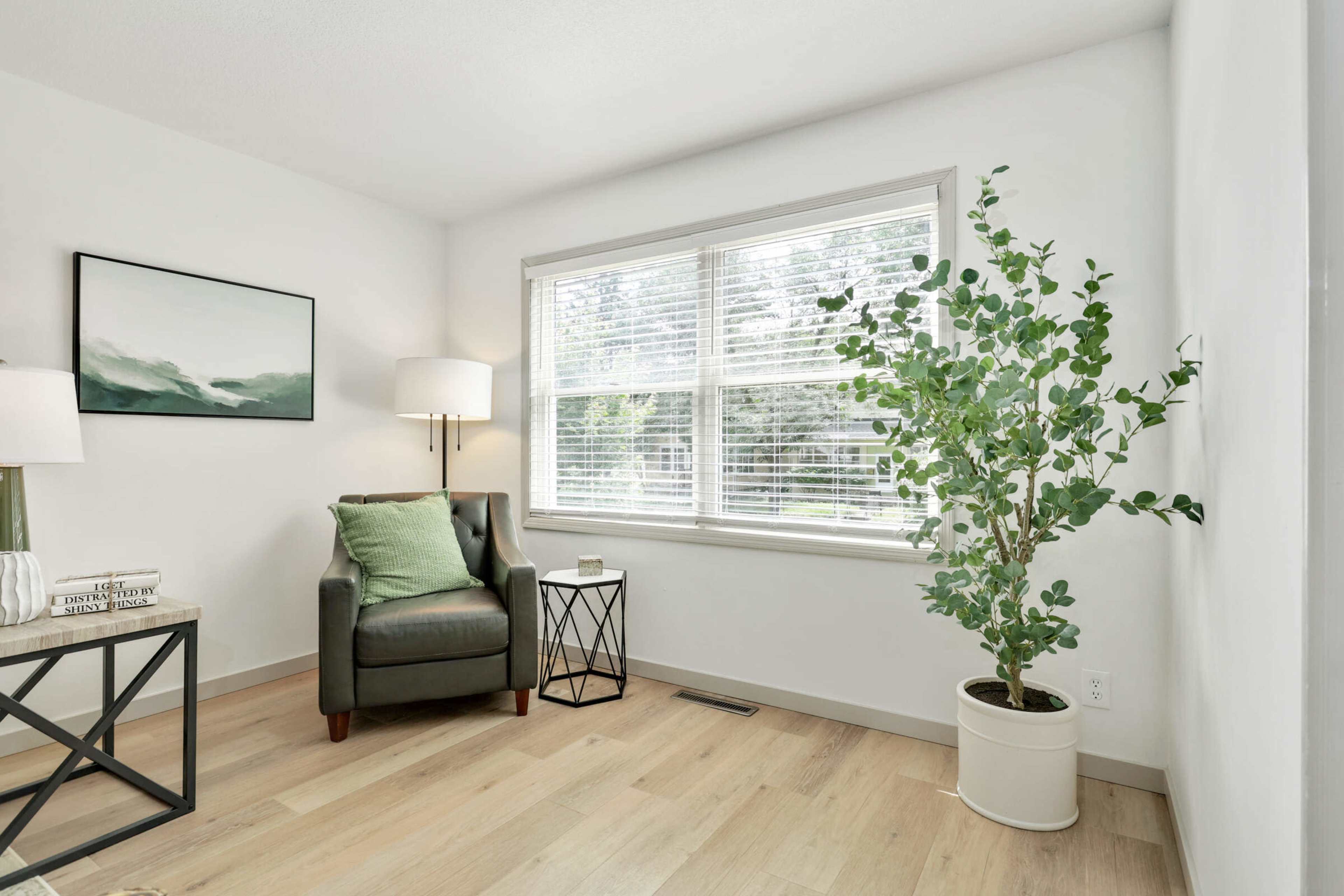 A cozy corner of a room features a green armchair beside a small black side table, a white planter with a plant, and a large window with blinds.